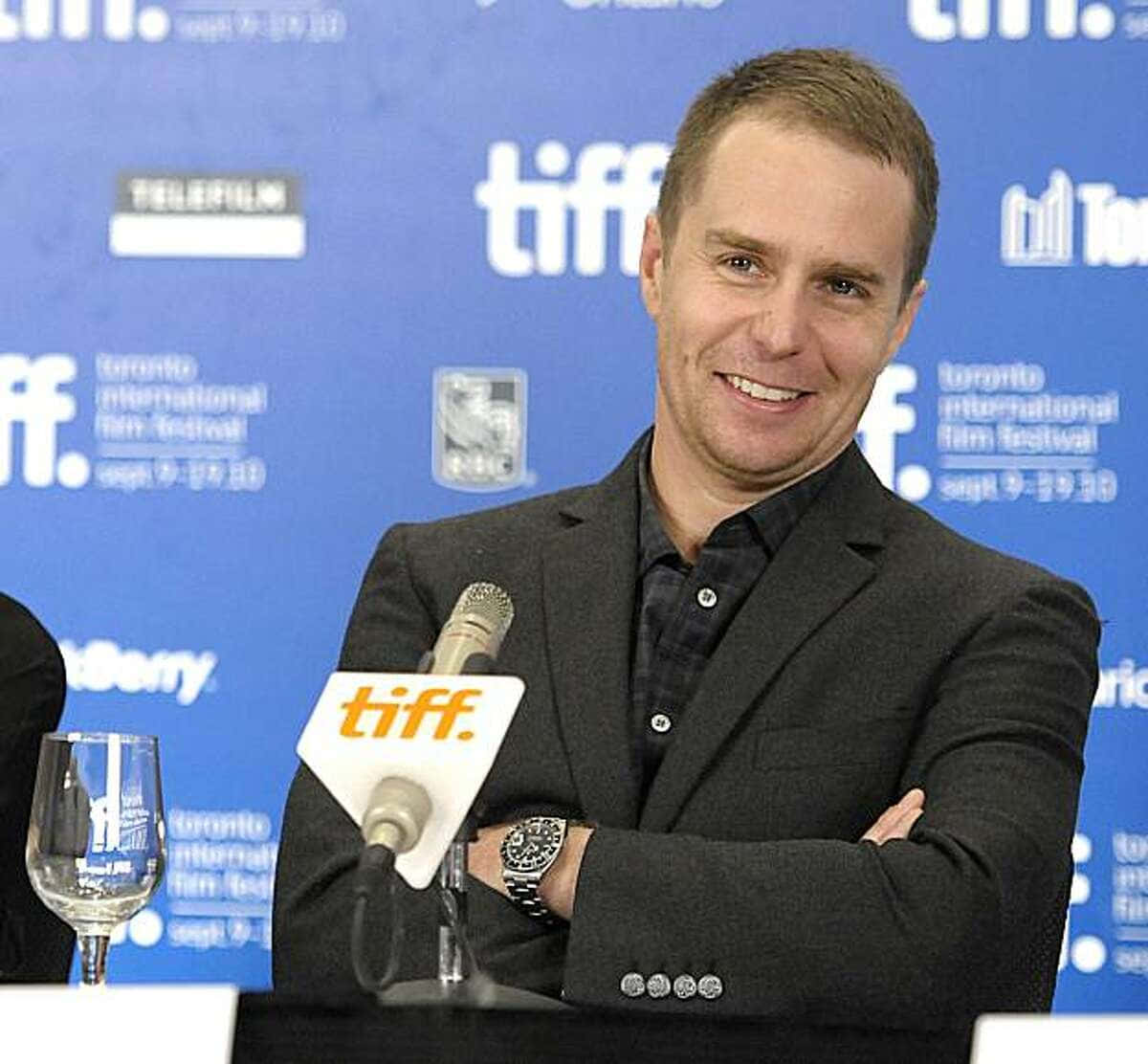 攝影 桌布 壁紙 — 一位身穿深色西裝、面帶微笑的男子自信地站在TIFF記者會上，雙臂交叉，面前放著麥克風。
