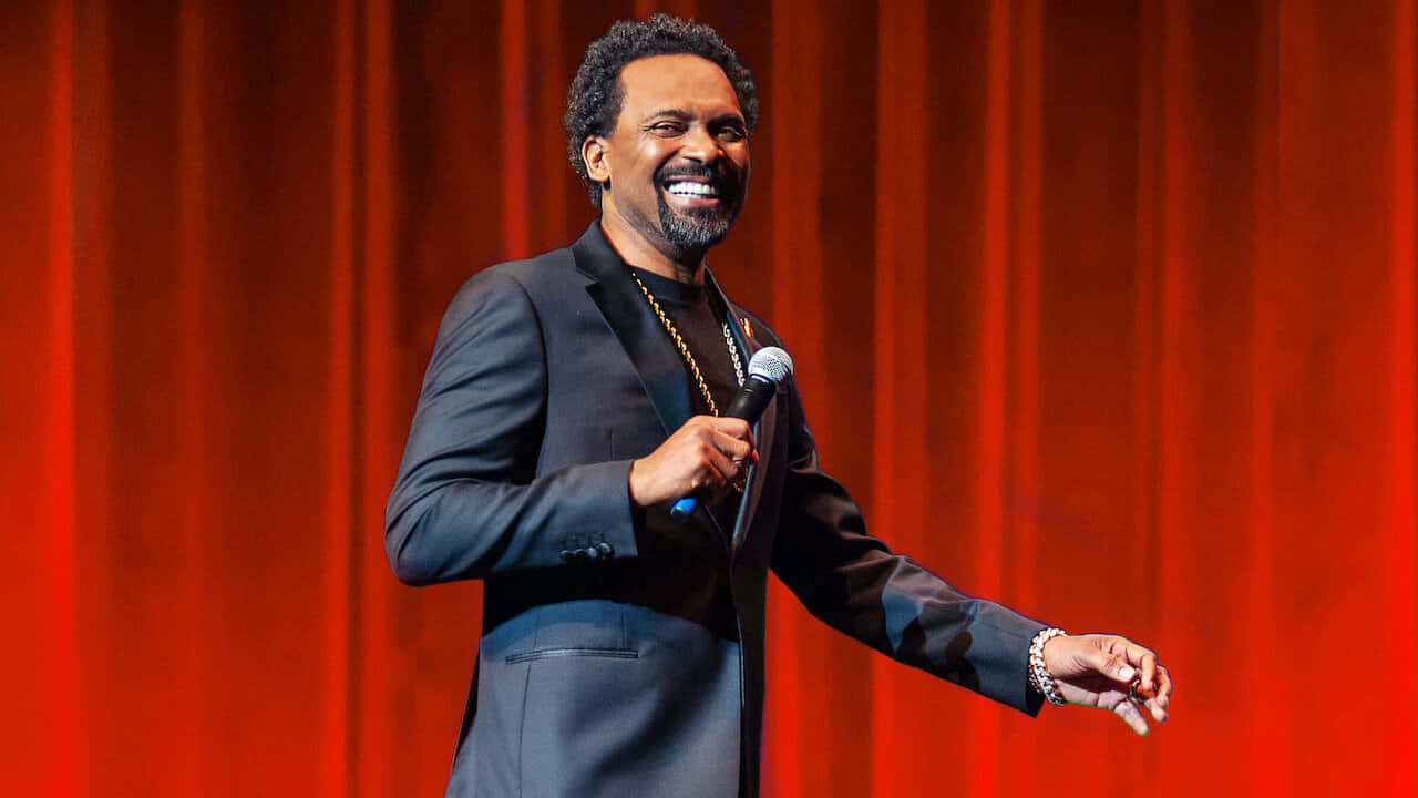 Mike Epps 桌布 壁紙 — 一位身穿深色西裝、面帶微笑的男子，在鮮紅色帷幕前的舞台上手持麥克風。