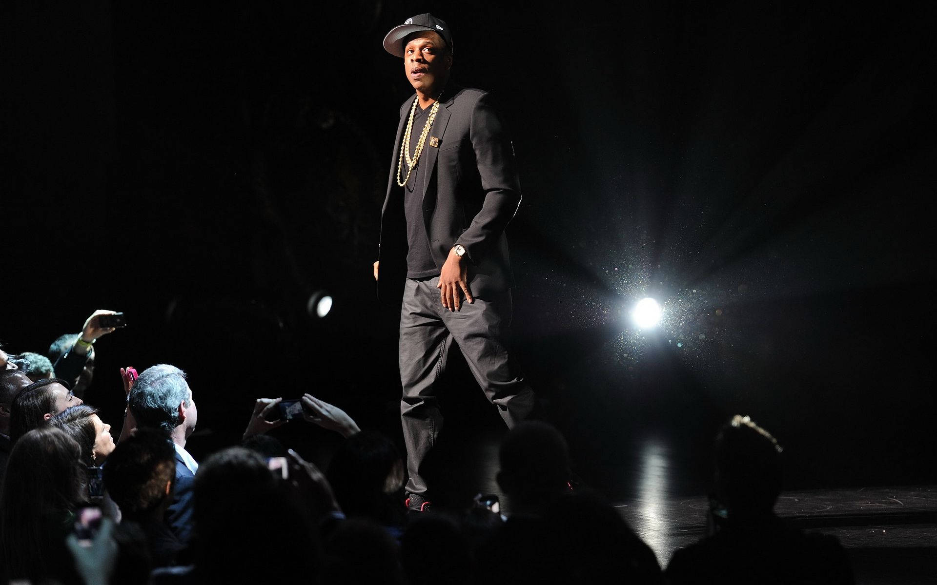 Jay-z 桌布 壁紙 — 一位身穿深色西裝和帽子的表演者站在舞台中央的聚光燈下，觀眾用手機記錄這一刻。