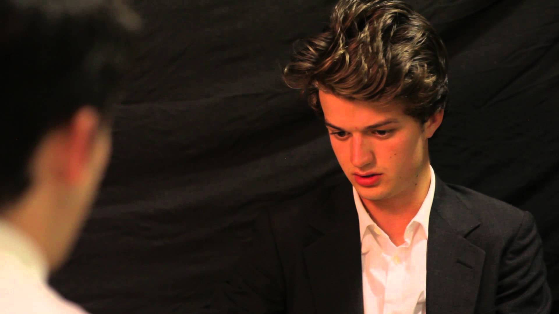 Joe Keery 桌布 壁紙 — 一位身穿深色西裝和白色襯衫的年輕男子在黑色背景下專注地向下看，而另一個人正面向他。