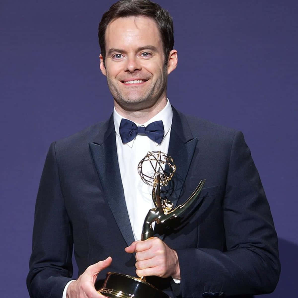 bill hader 桌布 壁紙 — 一位身穿深色西裝和領結的男士，在純紫色背景下自豪地手持金色艾美獎獎座。