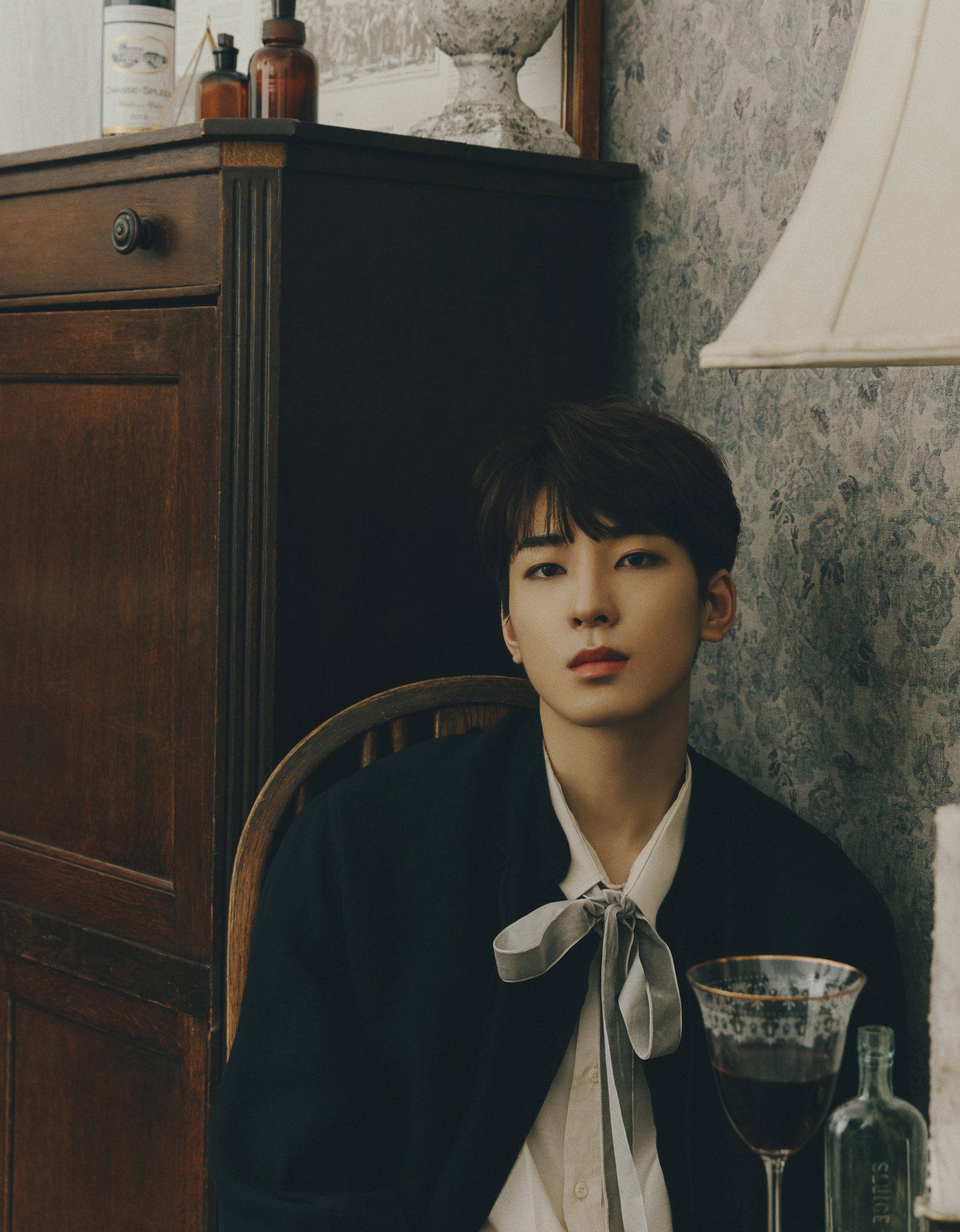 Wonwoo 桌布 壁紙 — 一位身穿深色西裝外套、白襯衫和灰色領結的年輕男子坐在木椅上，旁邊是復古櫥櫃和圖案壁紙。