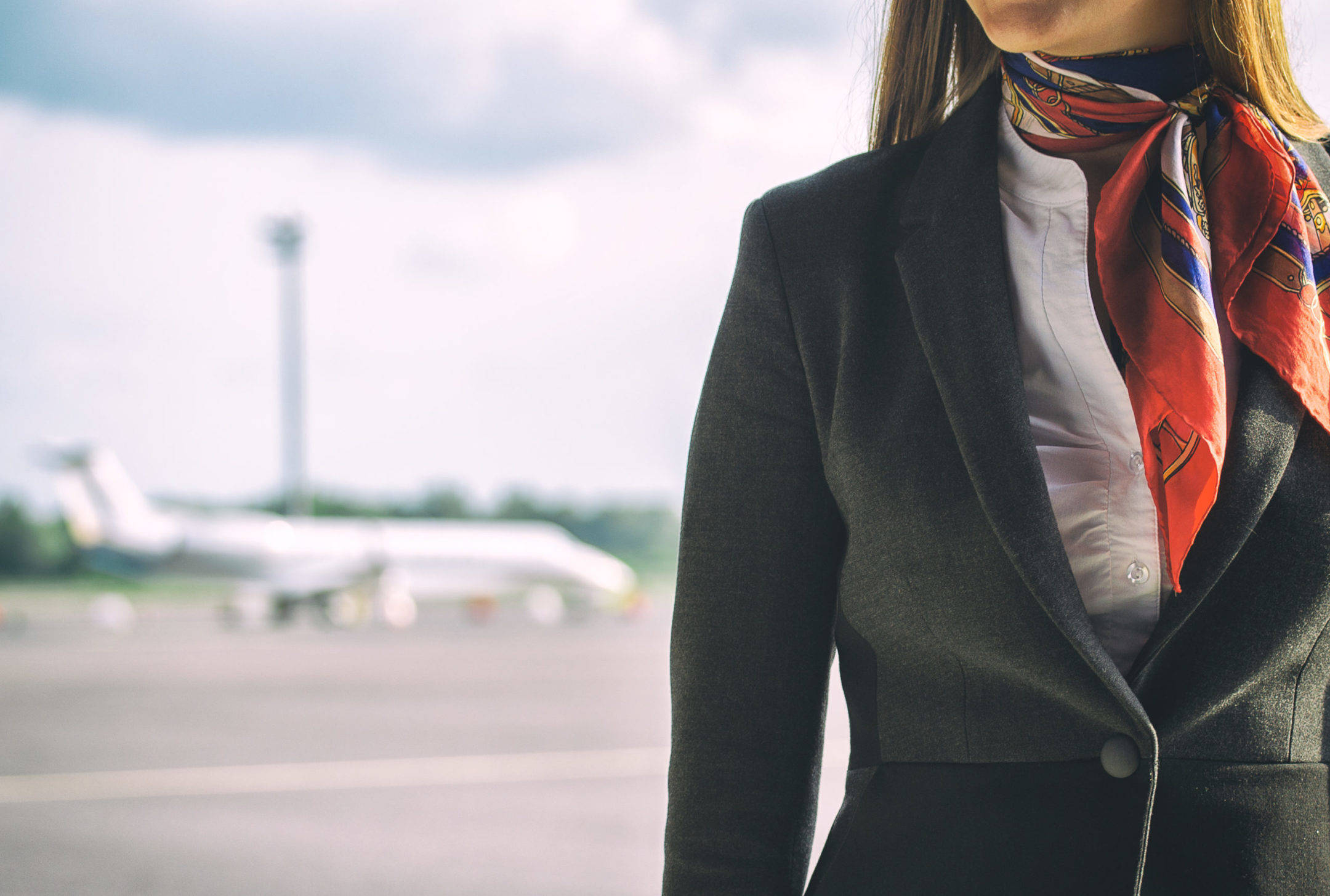 Airport 桌布 壁紙 — 一位身穿深色西裝外套和彩色圍巾的空中服務員自信地站在機場停機坪上，背景是模糊的飛機。
