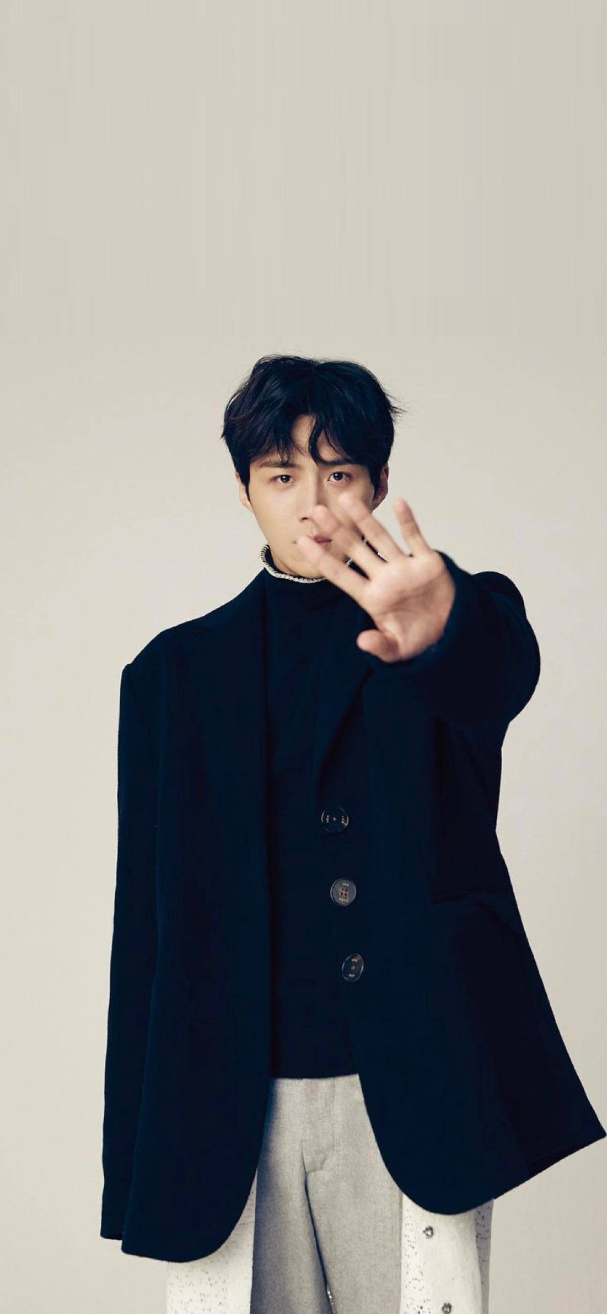 Kim Seon Ho 桌布 壁紙 — 一位身穿深色西裝外套和淺色褲子的年輕男子，在純米色背景前做出停止手勢，將手向前伸出。