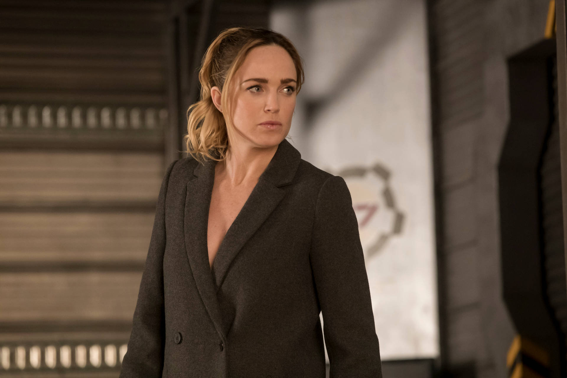 身穿深色西裝外套的女性在工業環境中表情嚴肅地站立。 — Caity Lotz 動圖 壁紙