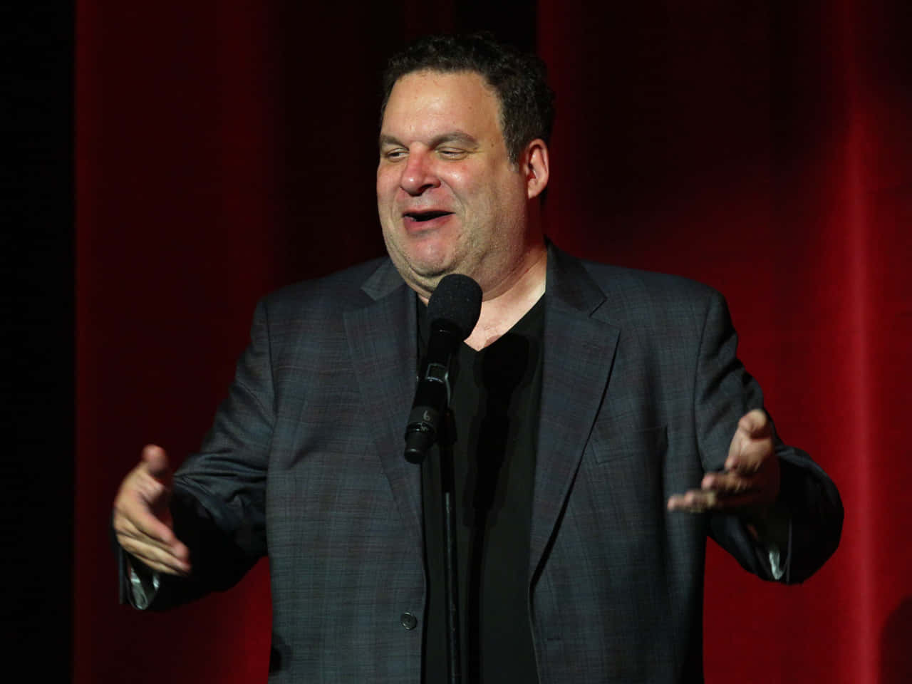 Jeff Garlin 桌布 壁紙 — 一位身穿深色西裝的喜劇演員在舞台上表演，身後有紅色窗簾，他開懷大笑。