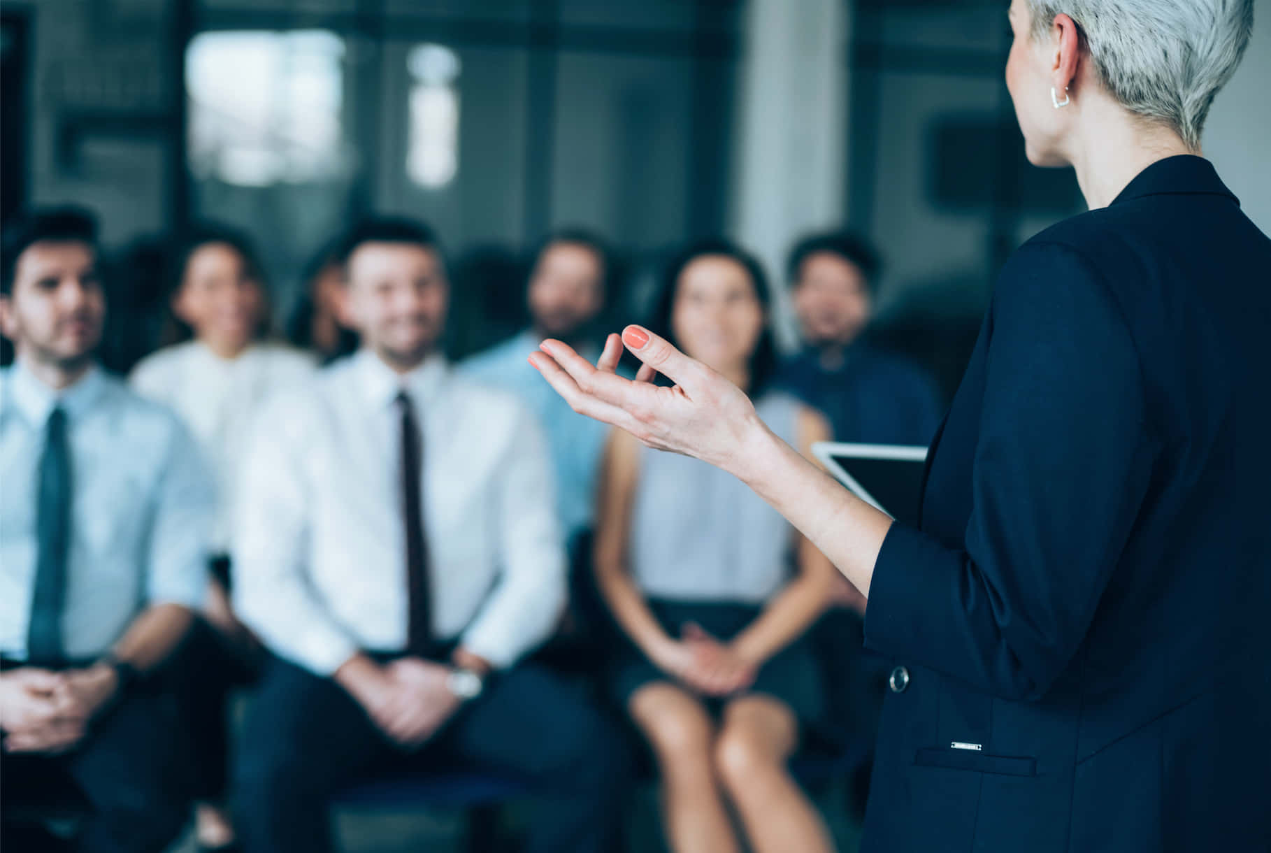 Leadership 桌布 壁紙 — 一位身穿深色西裝的女性在現代辦公室環境中向模糊的觀眾做手勢並進行簡報。