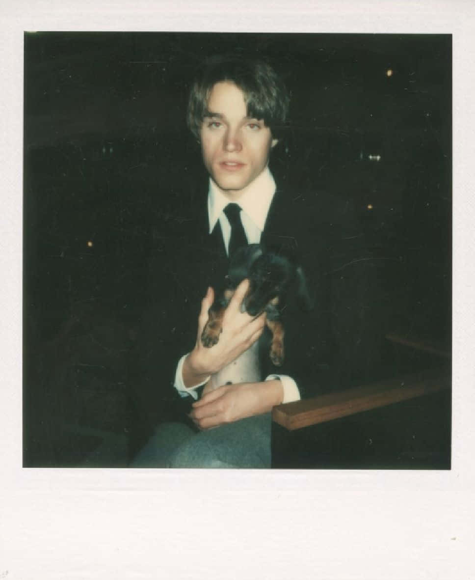 andy warhol 桌布 壁紙 — 一位身穿深色西裝的年輕男子在昏暗的房間裡抱著一隻臘腸犬幼犬，帶有拍立得底片美學。