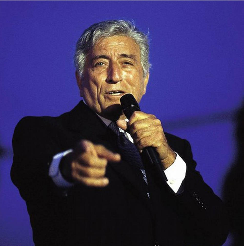 Singing 桌布 壁紙 — 一位身穿深色西裝的年長男性手持麥克風，在純藍色背景前向前指。