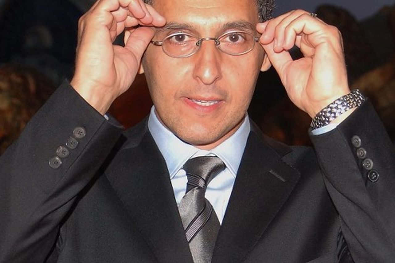 John Turturro 桌布 壁紙 — 一位身穿深色西裝的男子用雙手調整眼鏡，同時對著鏡頭微笑。