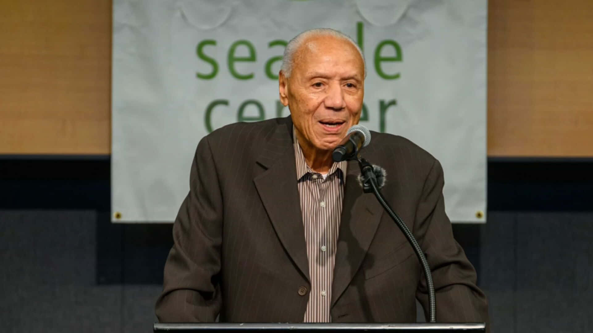 NBA 桌布 壁紙 — 一位身穿深色西裝的長者，在背後有「Seattle Center」橫幅的講台上對著麥克風講話。