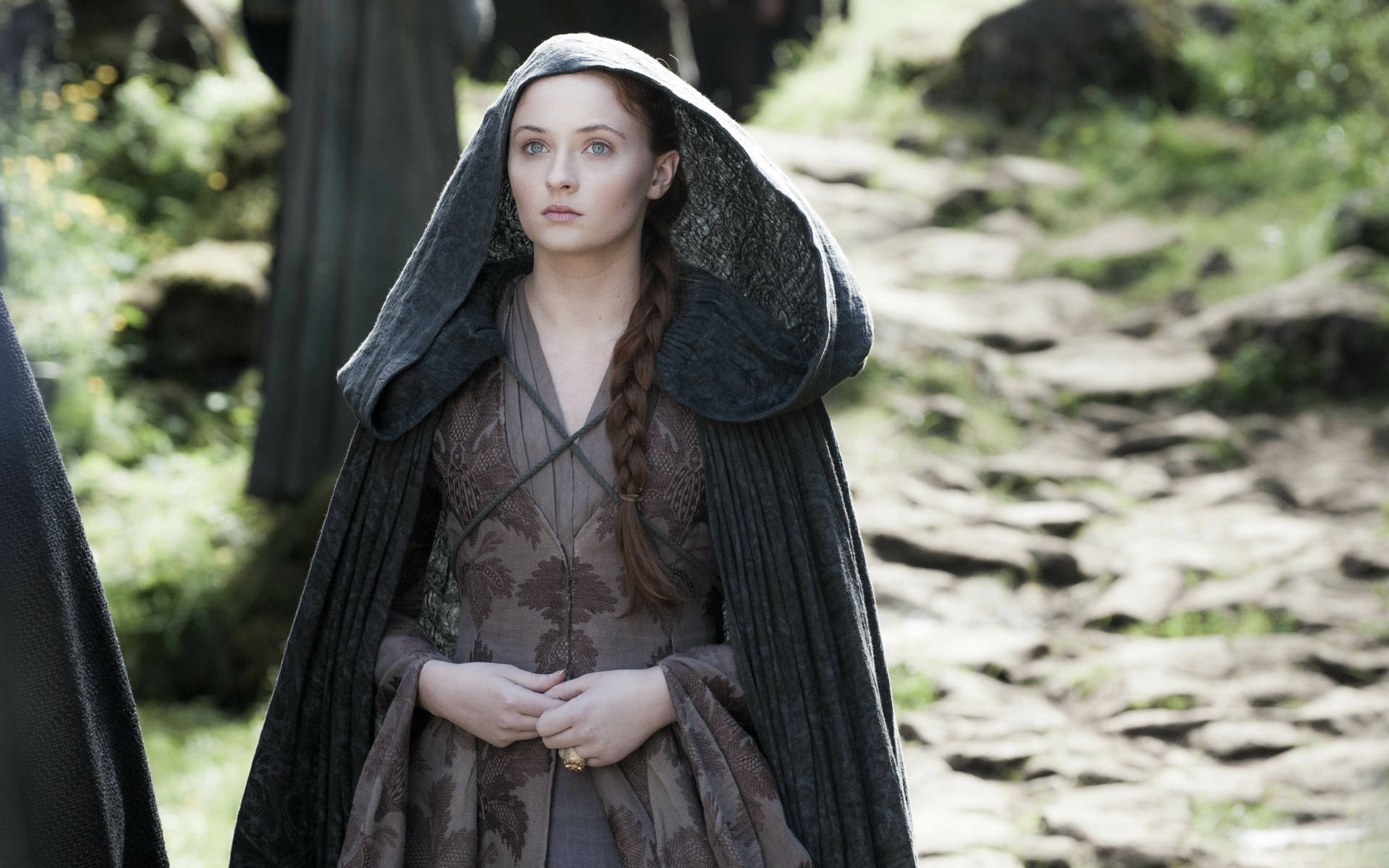 Sansa Stark 桌布 壁紙 — 一位身穿深色連帽斗篷的女子站在陽光普照的石徑上，穿越一片鬱鬱蔥蔥的綠色森林。
