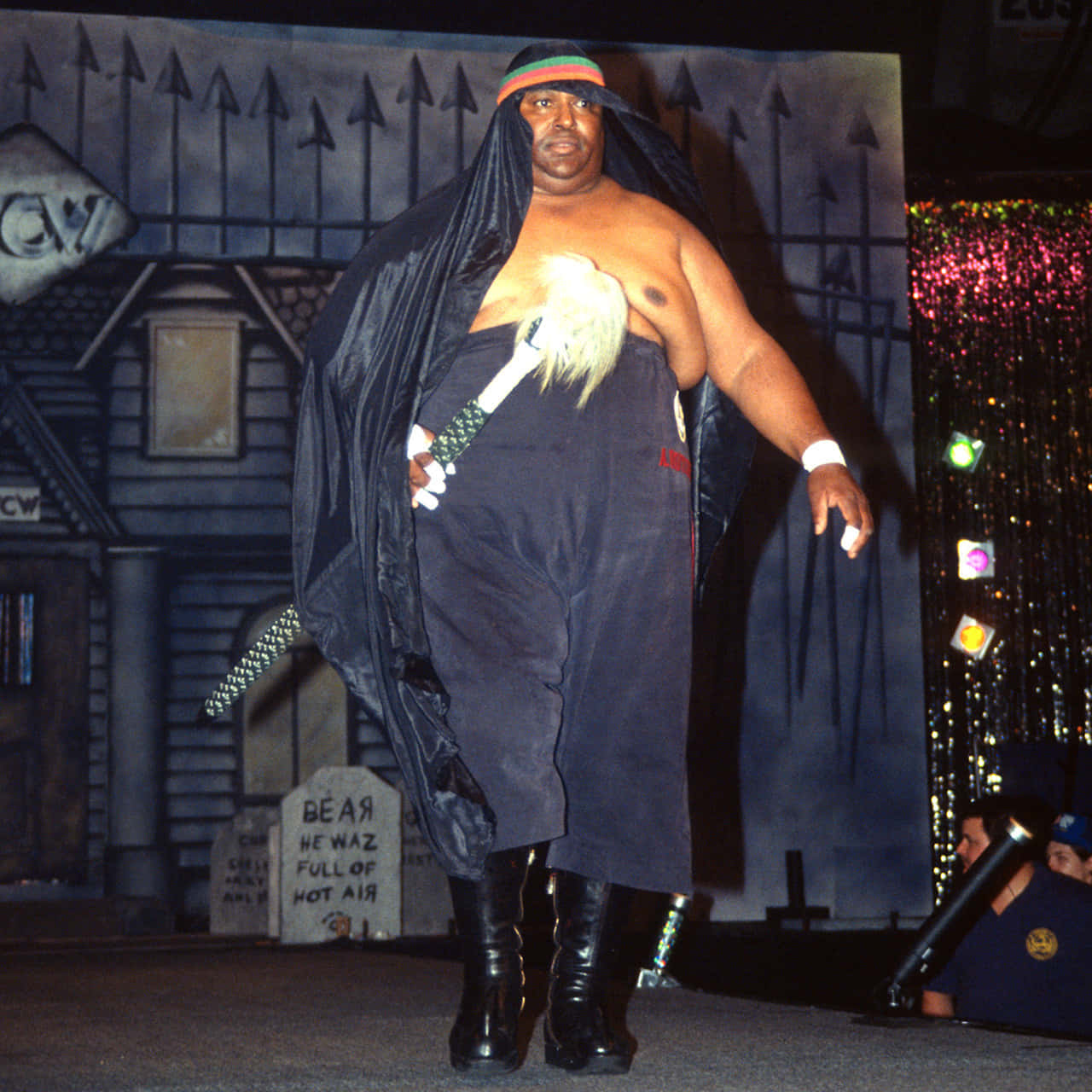 Abdullah The Butcher 桌布 壁紙 — 一位身穿深色連帽服裝、留著白鬍子、手持劍的摔角手，自信地走在以鬼屋為背景的舞台上。