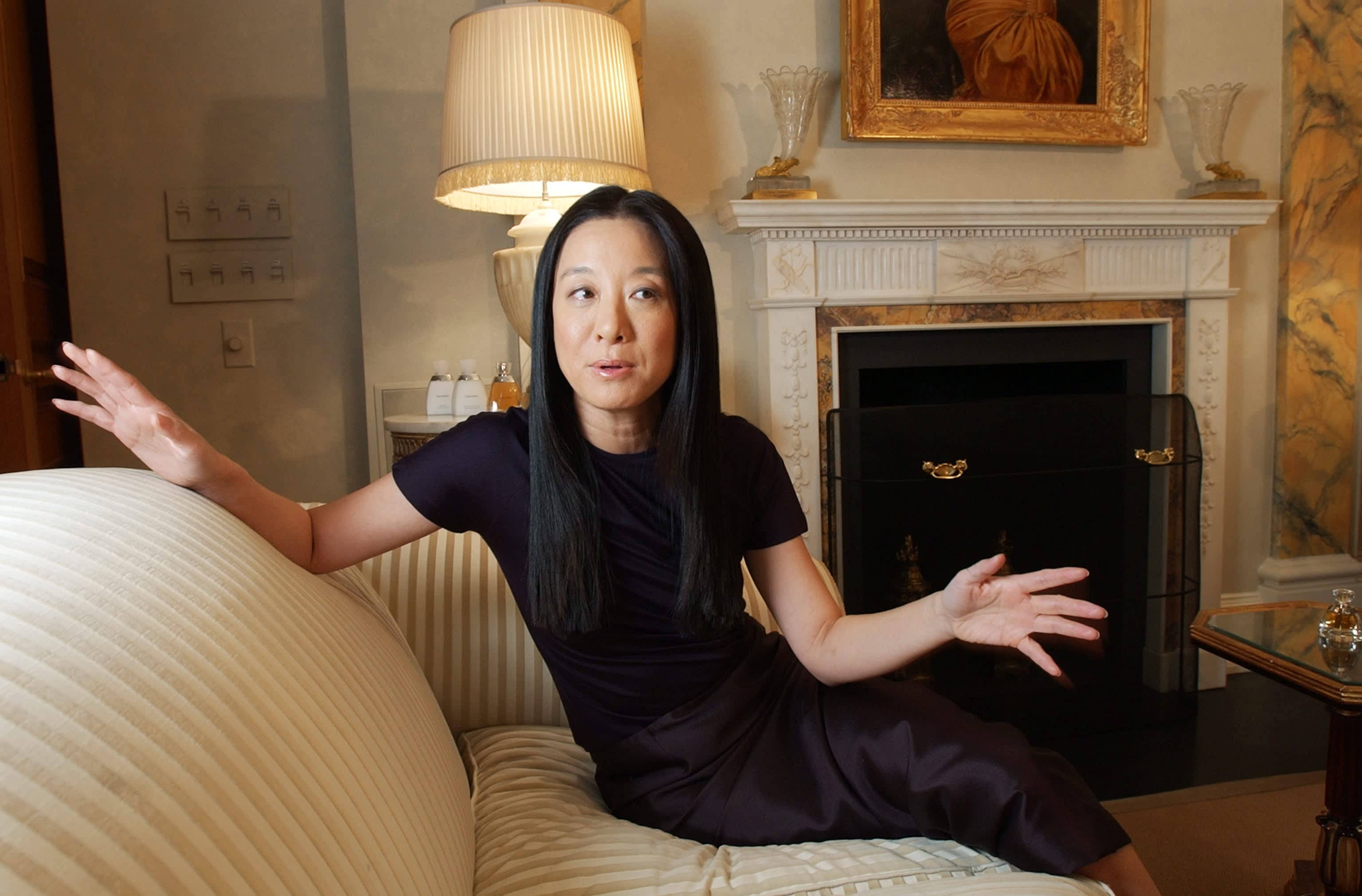 Asian Woman 桌布 壁紙 — 一位身穿深色連衣裙的女性放鬆地坐在條紋沙發上，身處一個溫暖照明、優雅的客廳，配有壁爐和華麗裝飾。