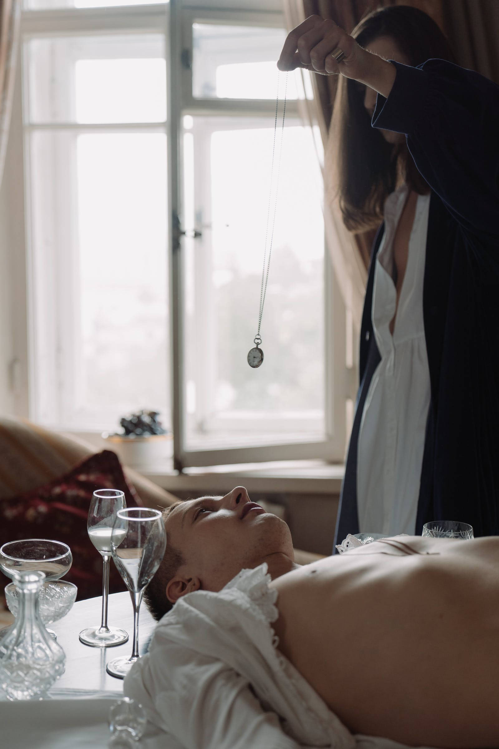 Hypnosis 桌布 壁紙 — 一位身穿深色長袍的女性在陽光充足的房間裡，手持懷錶放在一位斜倚的男子上方，房間裡有酒杯和窗景。
