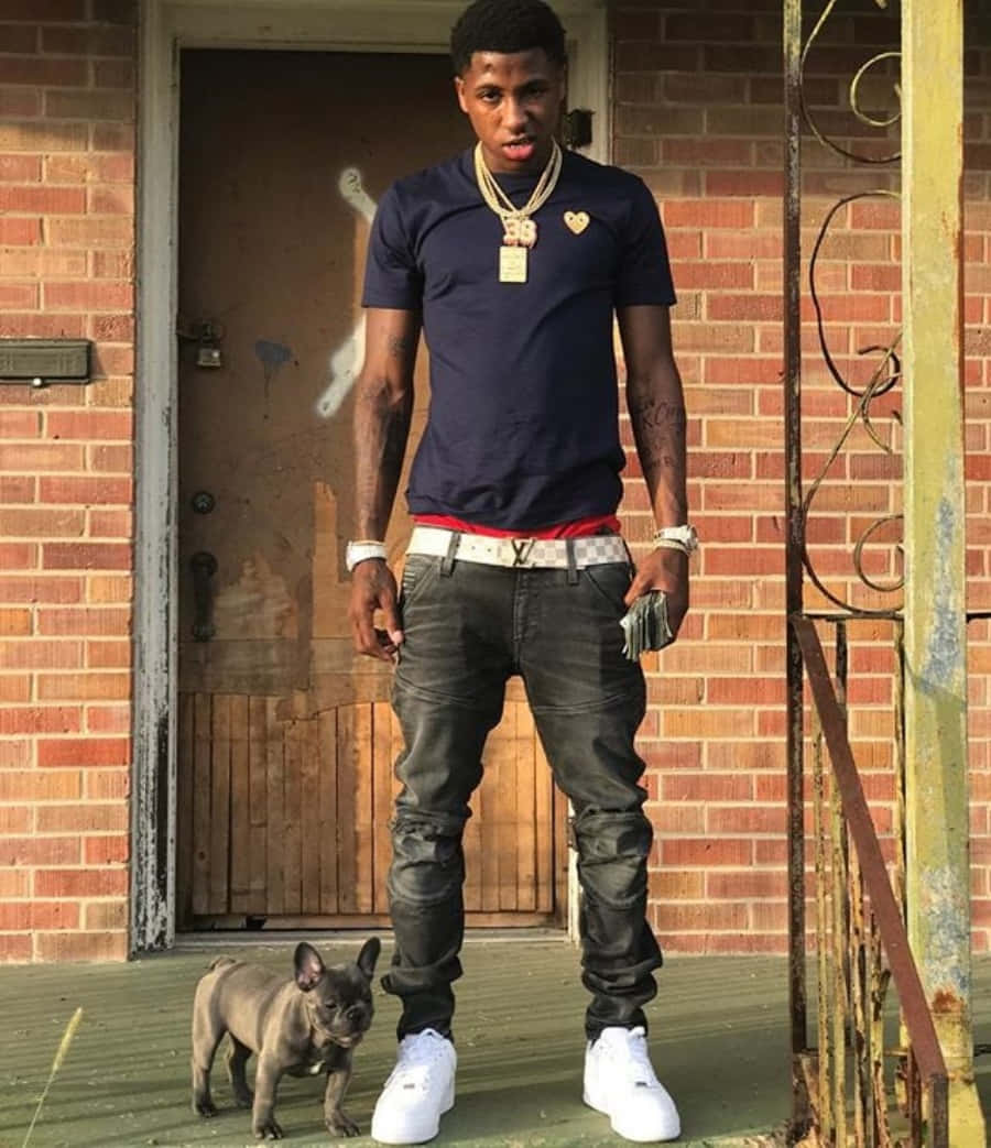 NBA YoungBoy 桌布 壁紙 — 一位身穿深藍色T恤和白色運動鞋的年輕男子自信地站在門廊上，身旁有一隻小灰色法國鬥牛犬。