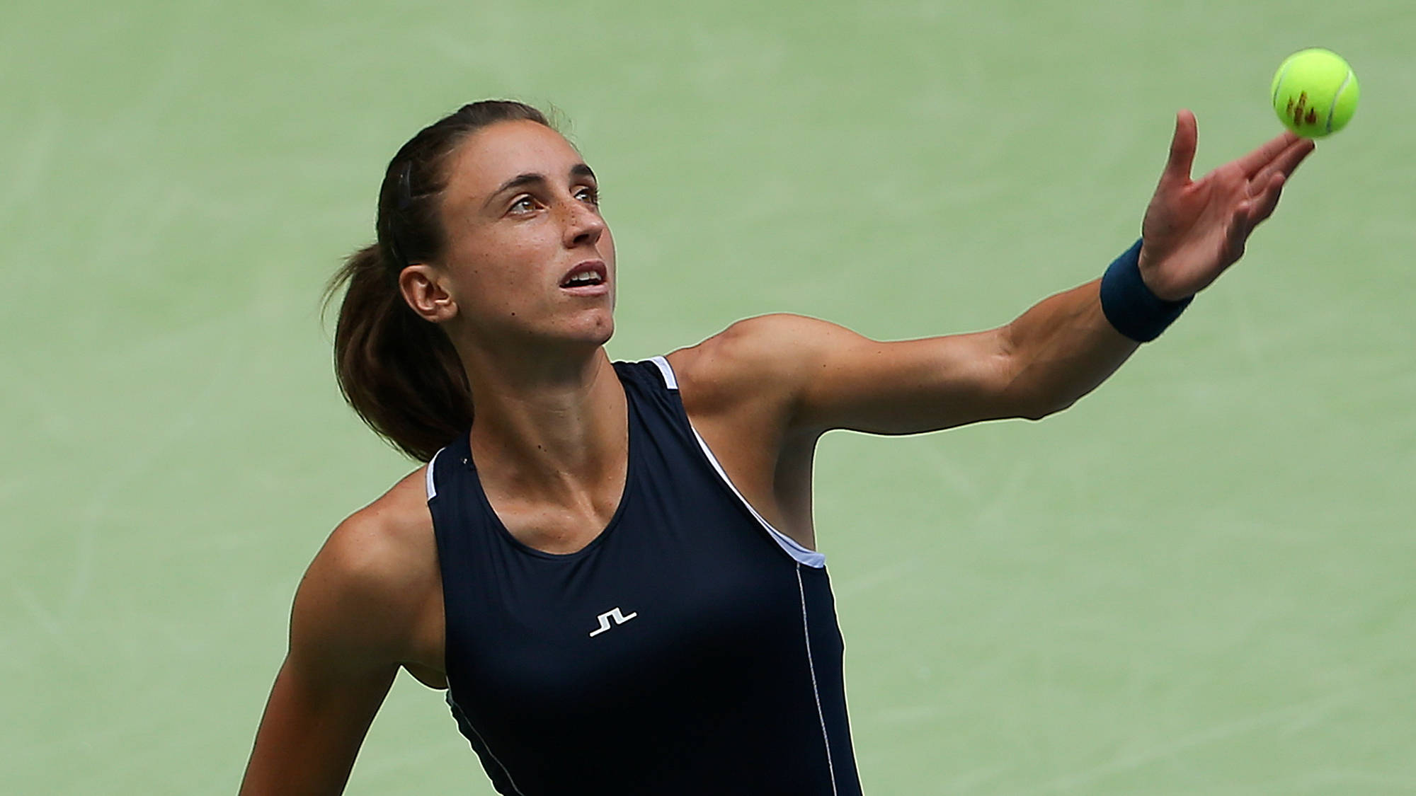 Athletic 桌布 壁紙 — 一位身穿深藍色上衣的女網球選手，在綠色球場背景下用右手將亮黃色的網球向上拋起。