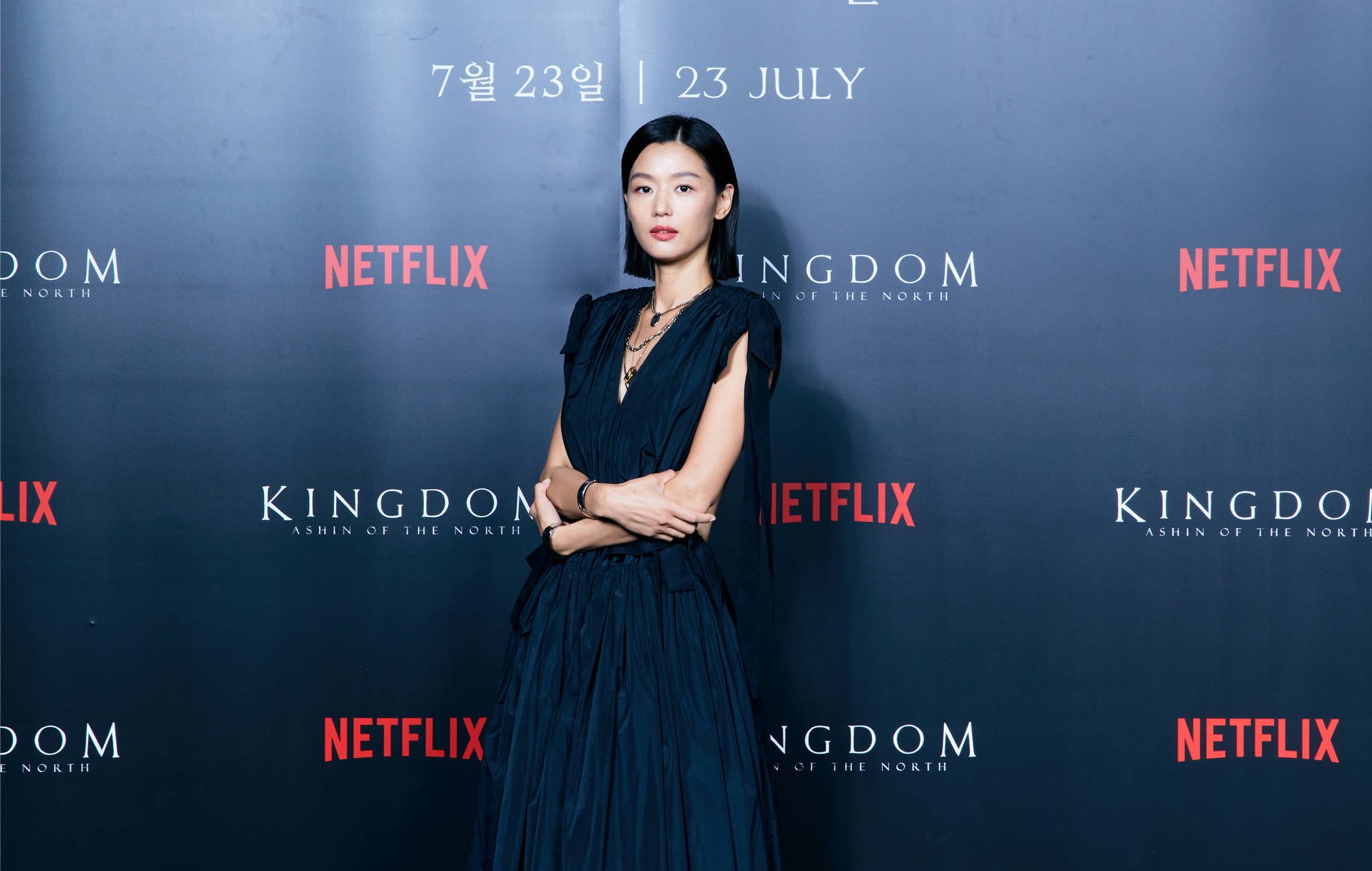 Jun Ji Hyun 桌布 壁紙 — 一位身穿深藍色禮服的優雅女士自信地站在Netflix Kingdom活動現場，身後有宣傳文字。