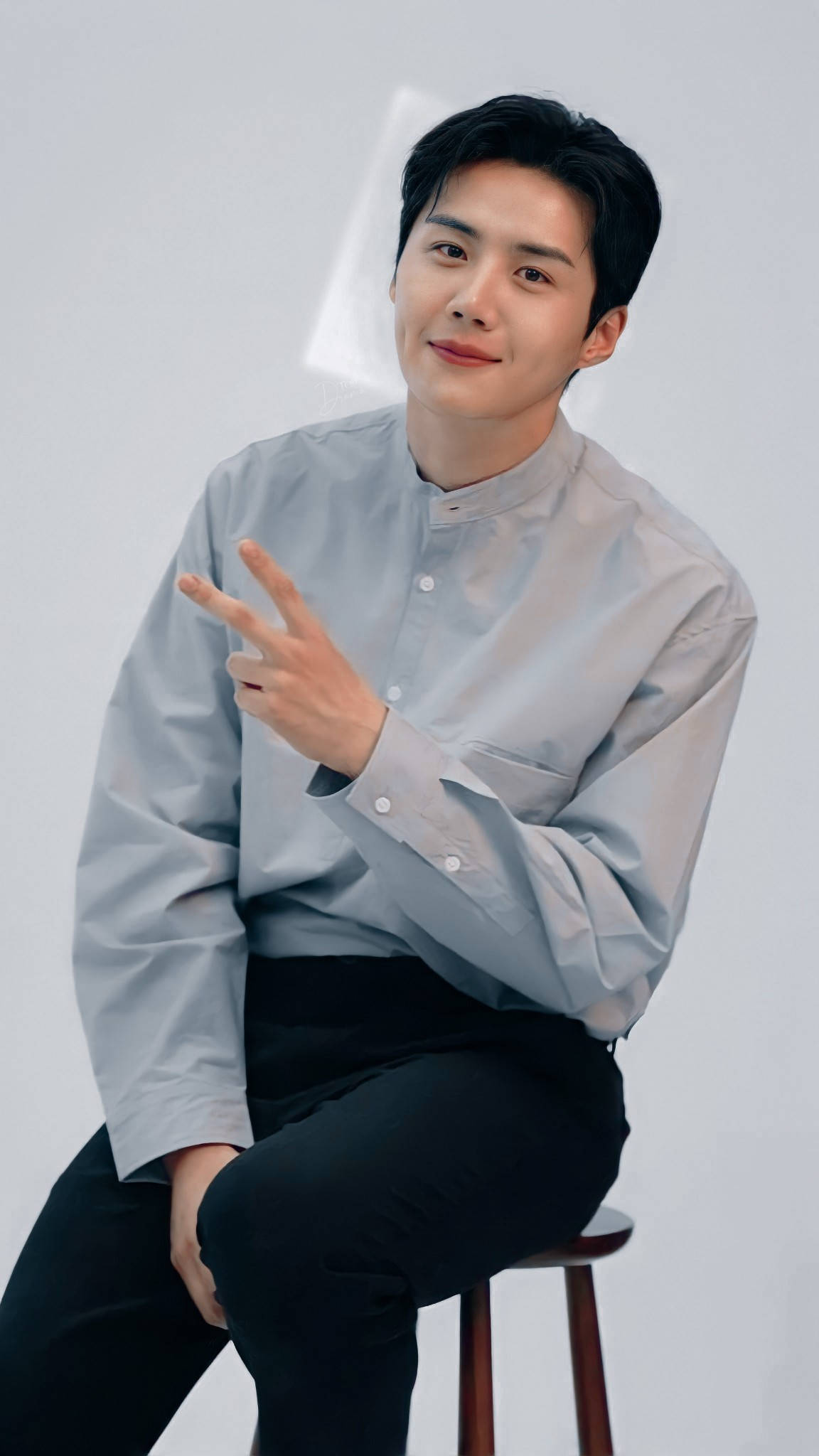 Kim Seon Ho 桌布 壁紙 — 一位身穿淺灰色企領襯衫的年輕男子坐在凳子上，在純白色背景前做出和平手勢。