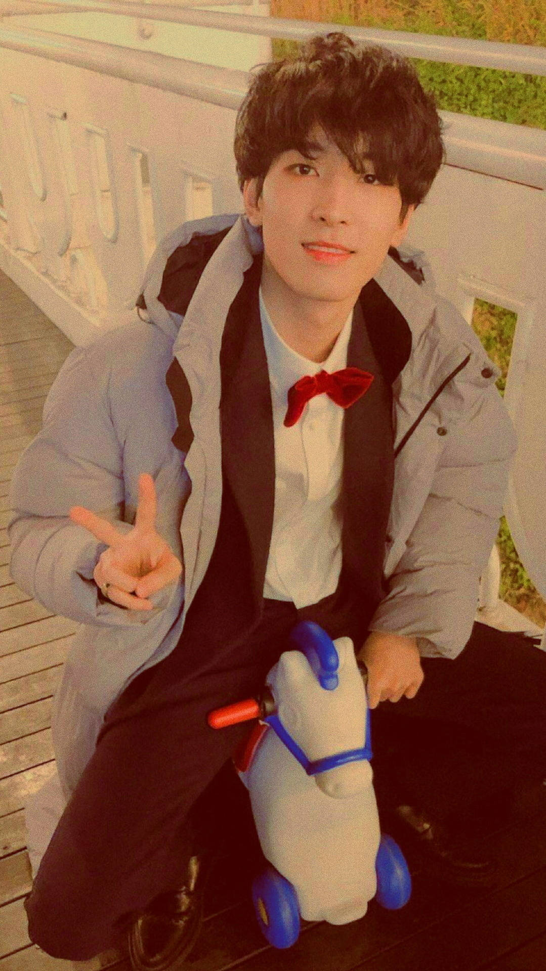 Wonwoo 桌布 壁紙 — 一位身穿淺灰色羽絨外套和紅色領結的年輕男子微笑著坐在木甲板上，旁邊是一隻白色和藍色的玩具馬。