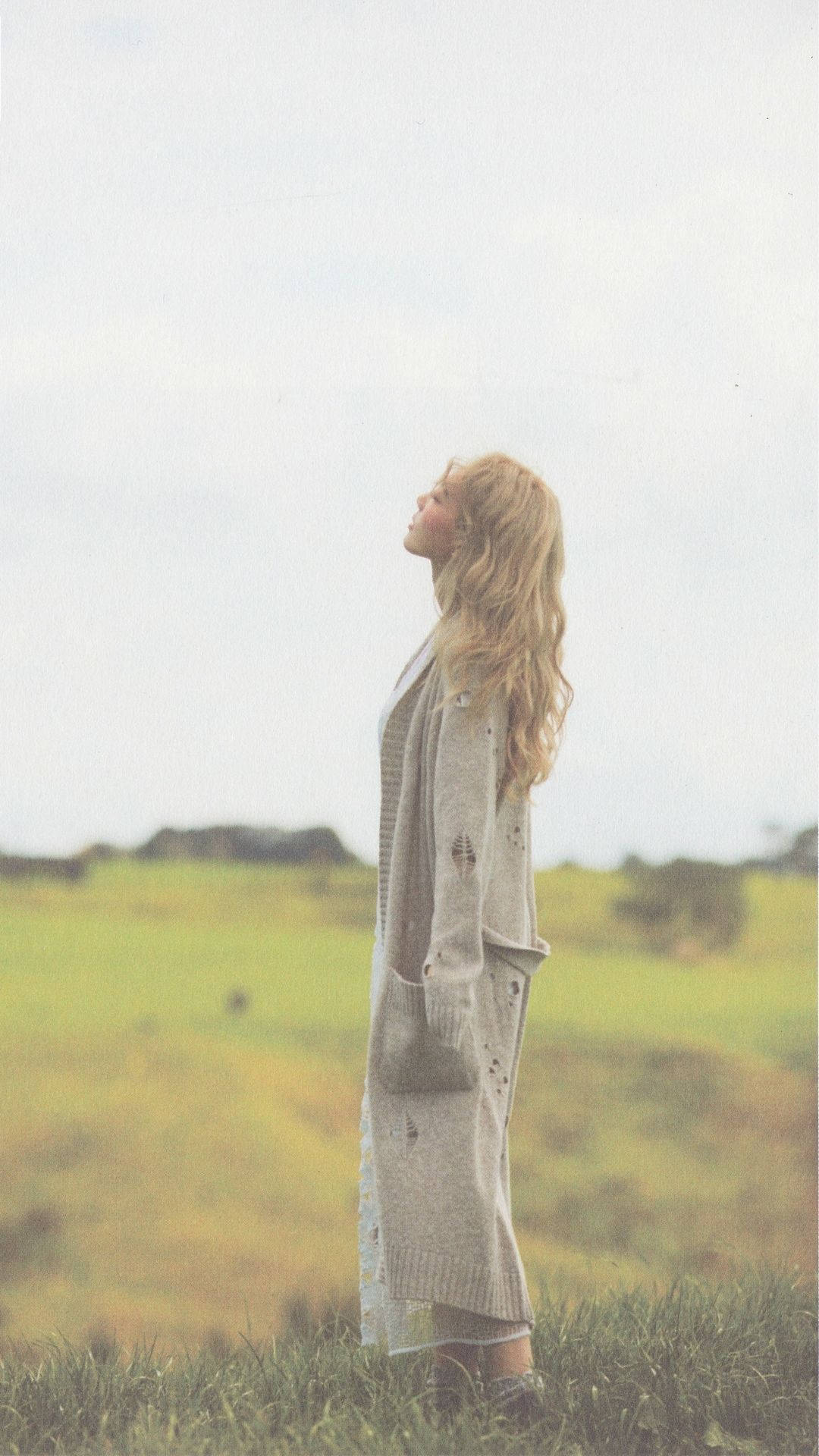 Taeyeon 桌布 壁紙 — 一位身穿淺灰色開襟毛衣的女性站在草地上，頭向後仰，閉上眼睛，沐浴在柔和的多雲光線中。