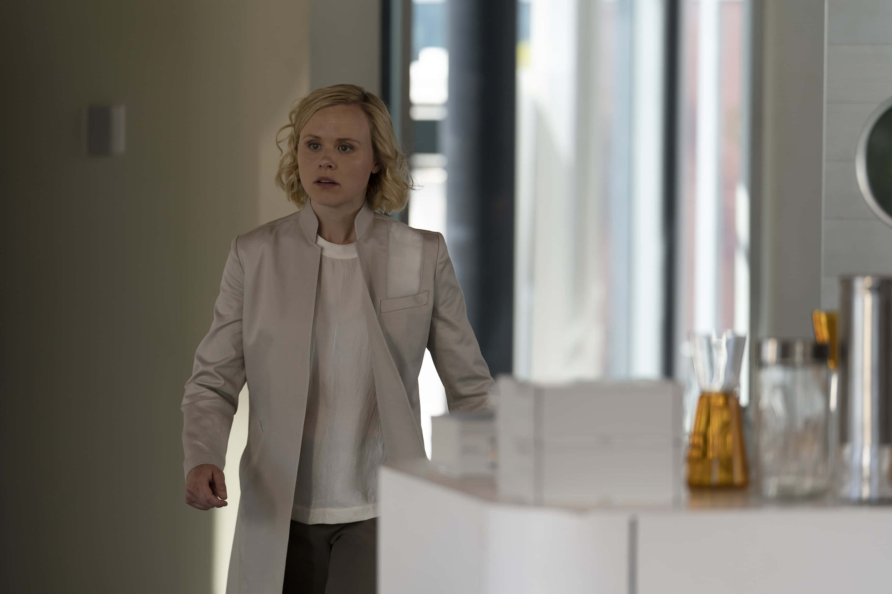 Alison Pill 桌布 壁紙 — 一位身穿淺米色外套的女性站在現代廚房裡，表情驚訝。