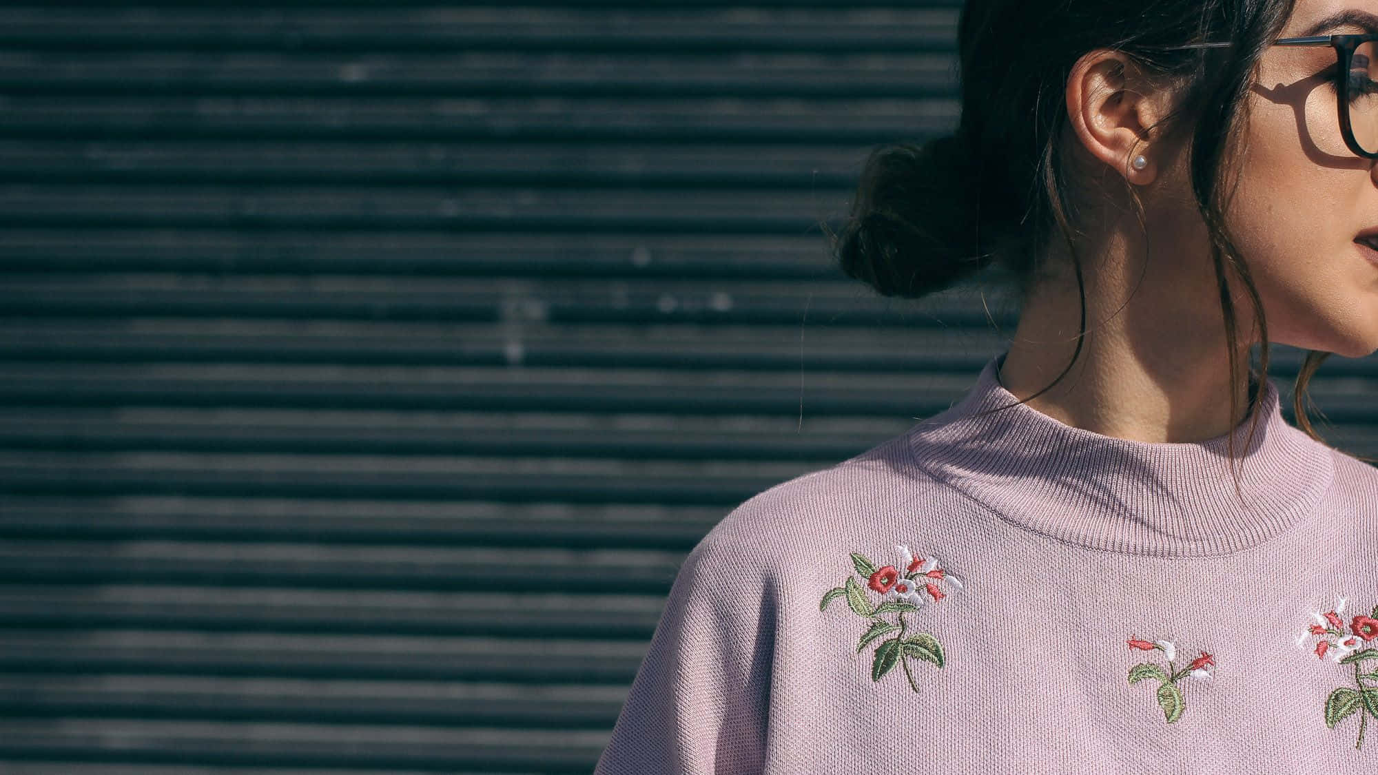 Girl Aesthetic Desktop 桌布 壁紙 — 一位身穿淺紫色刺繡花卉毛衣的女性，在明亮的陽光下站在深色波紋金屬牆前。
