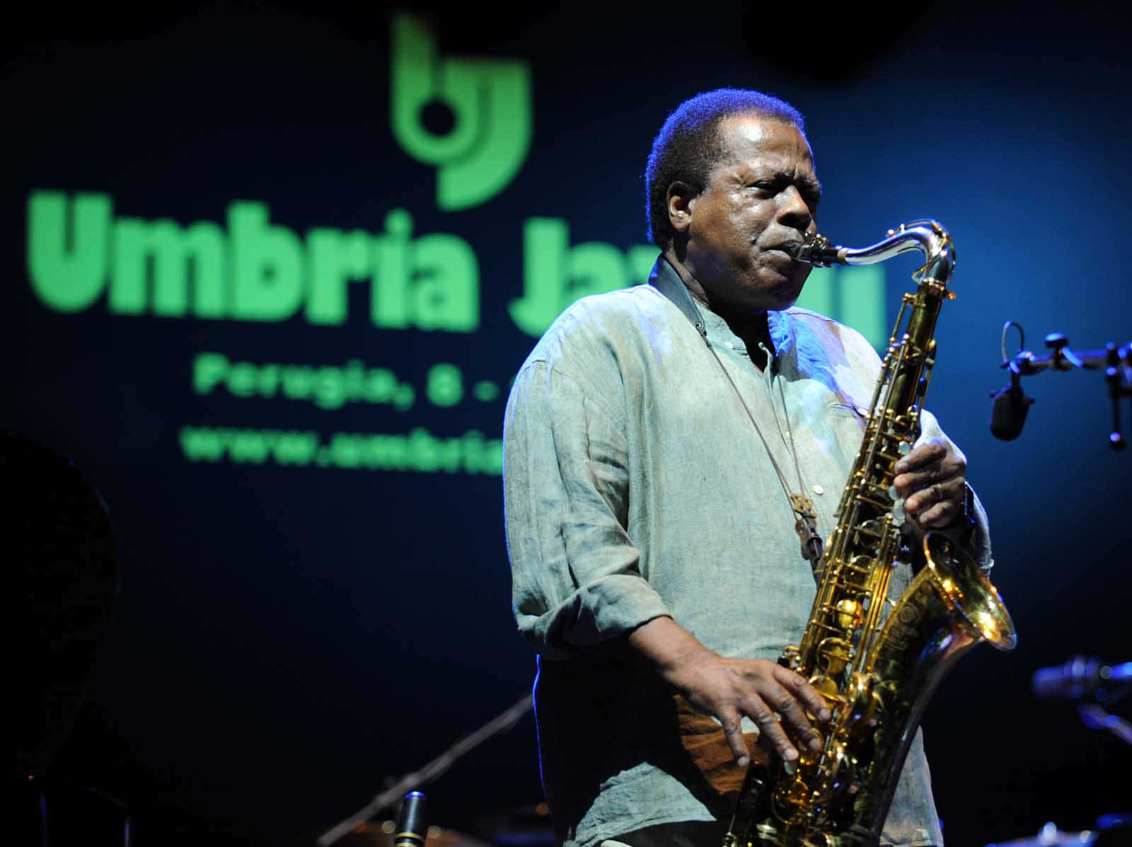 Wayne Shorter 桌布 壁紙 — 一位身穿淺色襯衫的爵士薩克斯風手在翁布里亞爵士音樂節的舞台燈光下熱情地演奏金色薩克斯風。