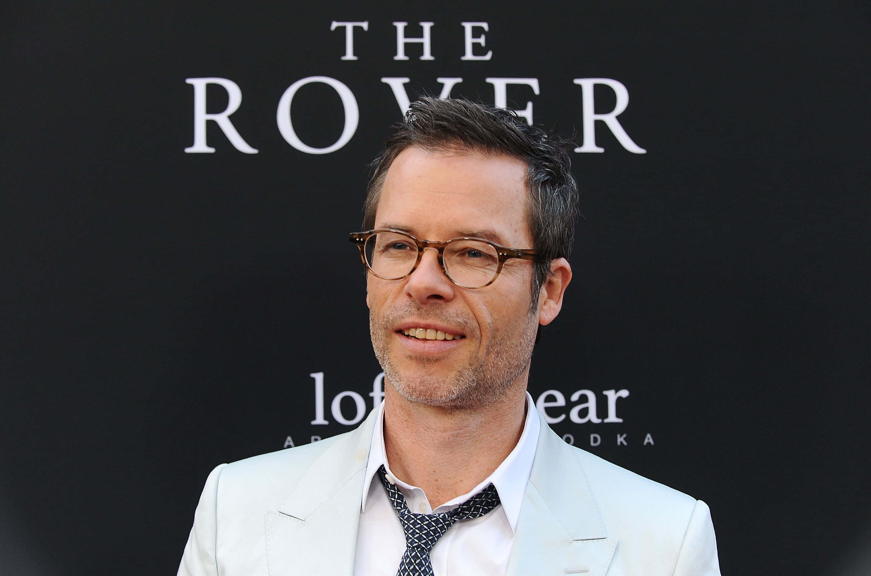 Guy Pearce 桌布 壁紙 — 一位身穿淺色西裝、戴眼鏡的男子在黑色背景前對著鏡頭微笑，背景上有白色文字。