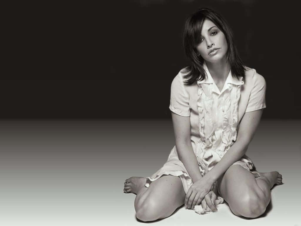 Gina Gershon 桌布 壁紙 — 一位身穿淺色連衣裙的女子赤腳坐在漸變地板上，對著黑暗的背景若有所思地凝視。