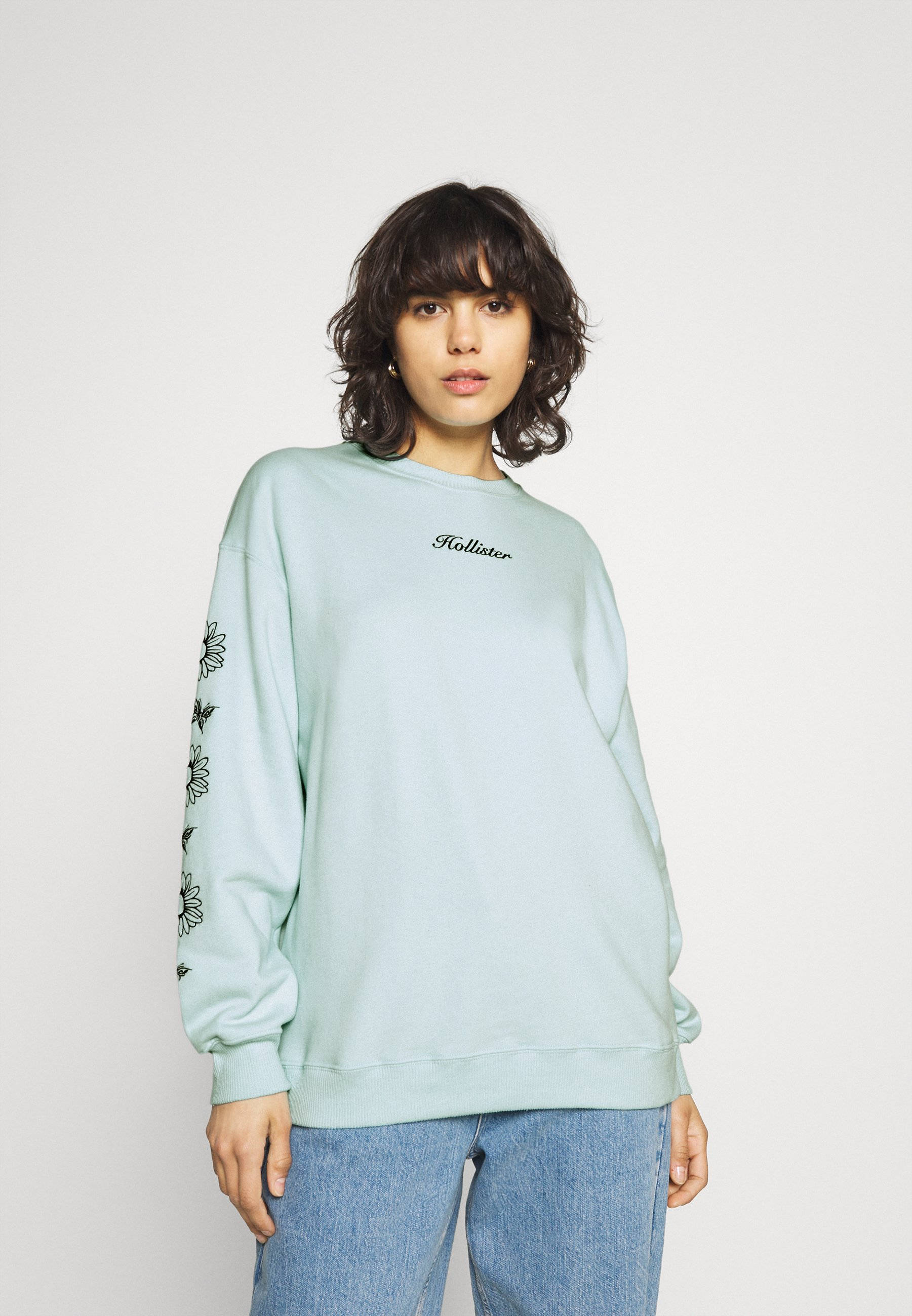 Hollister 桌布 壁紙 — 一位身穿淺藍色Hollister運動衫、袖子上有花卉刺繡的女性，站在純白色背景前。