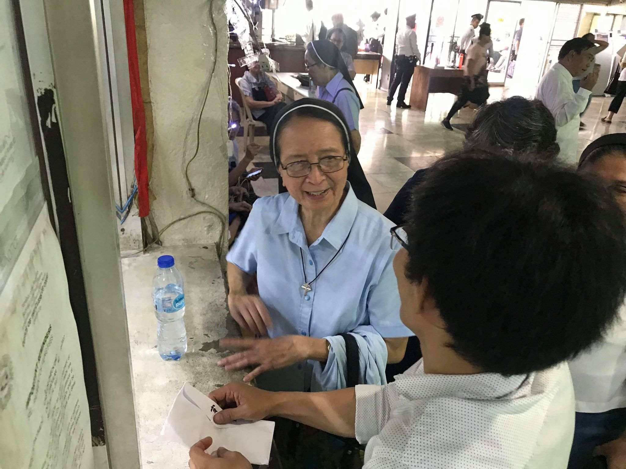 Smile 桌布 壁紙 — 一位身穿淺藍色修女服的微笑修女在繁忙的室內社區中心與訪客熱情互動。