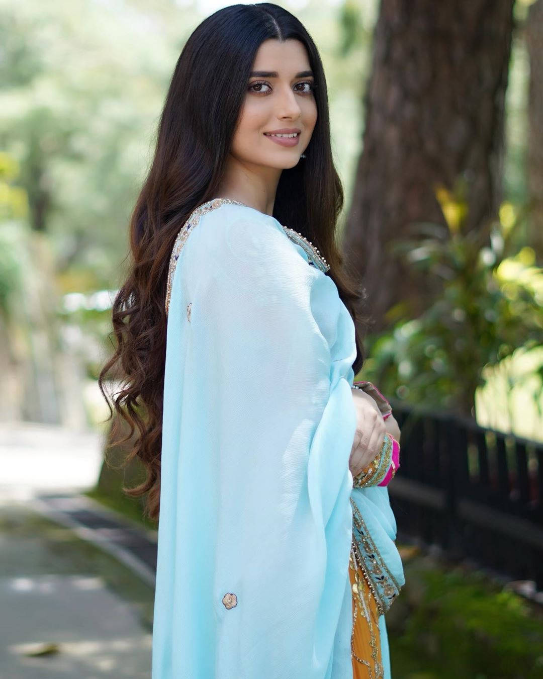 Indian 桌布 壁紙 — 一位身穿淺藍色傳統服飾的女性，在茂密的綠樹和斑駁的陽光中回頭微笑。