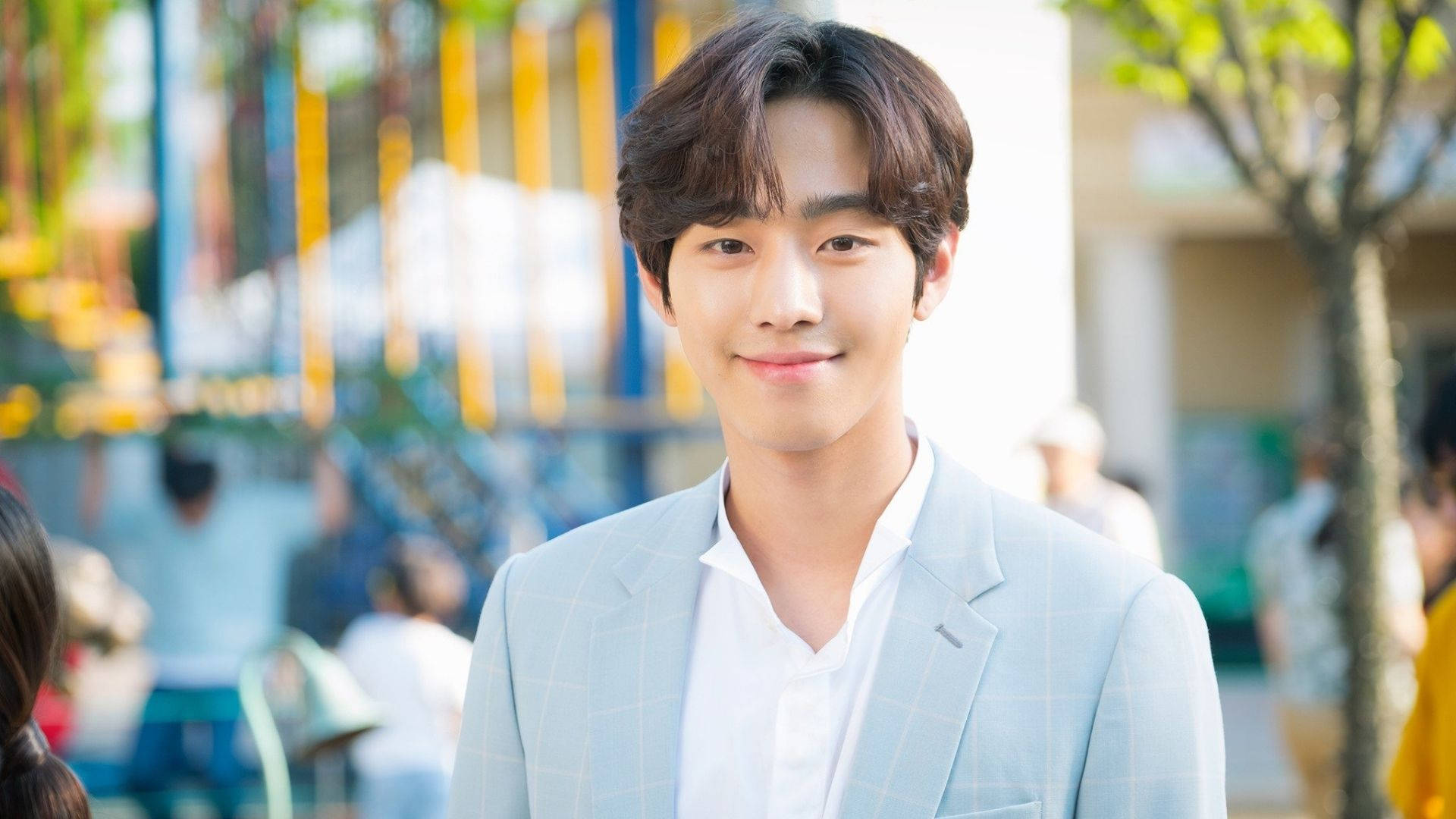 Ahn Hyo Seop 桌布 壁紙 — 一位身穿淺藍色格紋西裝的年輕男士，在明亮的戶外城市背景下對著鏡頭溫暖微笑。