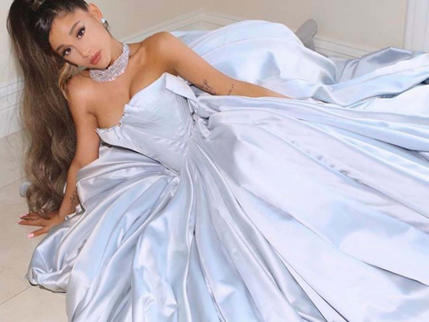 Ariana Grande Aesthetic 桌布 壁紙 — 一位身穿淺藍色緞面寬鬆長裙的女性優雅地斜倚在瓷磚地板上，佩戴著閃閃發光的頸鏈和一頭長波浪髮。