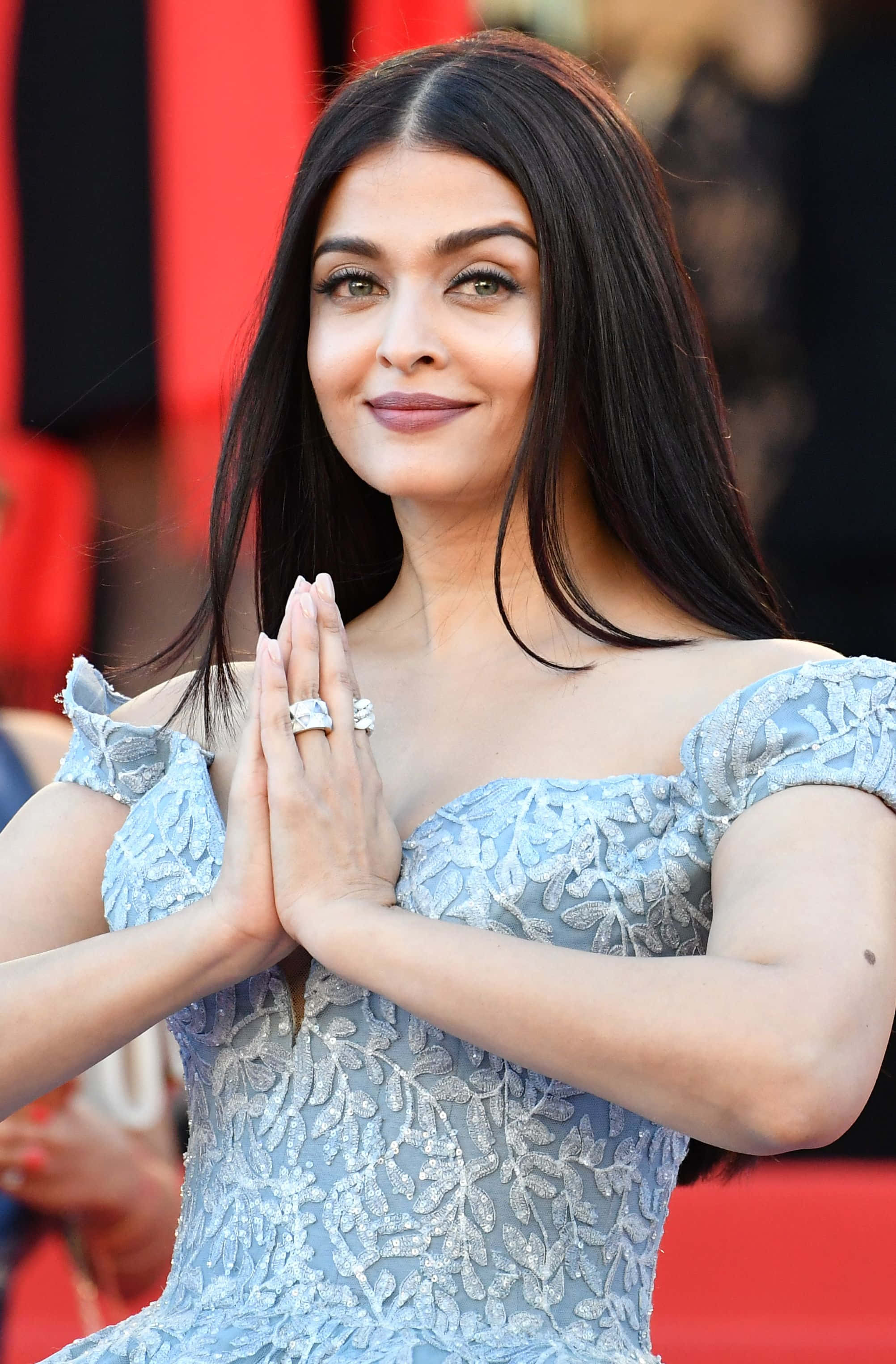 Aishwarya Rai 桌布 壁紙 — 一位身穿淺藍色蕾絲連身裙的女性，在紅地毯上雙手合十做出問候手勢，露出溫暖的笑容。