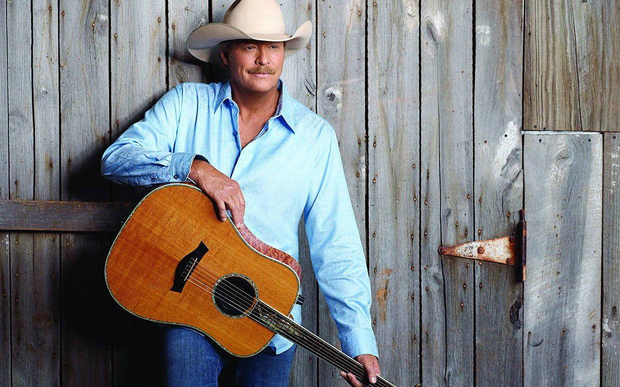 Alan Jackson 桌布 壁紙 — 一位身穿淺藍色襯衫、戴帽子的牛仔靠著風化的木頭，手持一把原聲吉他。