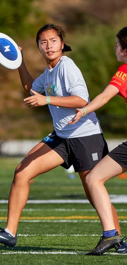 Android Ultimate Frisbee 桌布 壁紙 — 一位身穿淺藍色襯衫和黑色短褲的女運動員準備投擲一個帶有藍色星星的白色飛盤，同時在陽光明媚的綠色場地上被另一名球員防守。