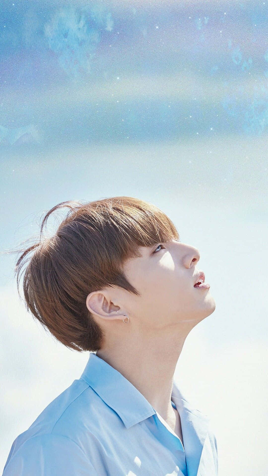 Jungkook 桌布 壁紙 — 一位身穿淺藍色襯衫的年輕男子仰望夢幻般的天空，天空佈滿柔軟的雲朵和閃爍的粒子。