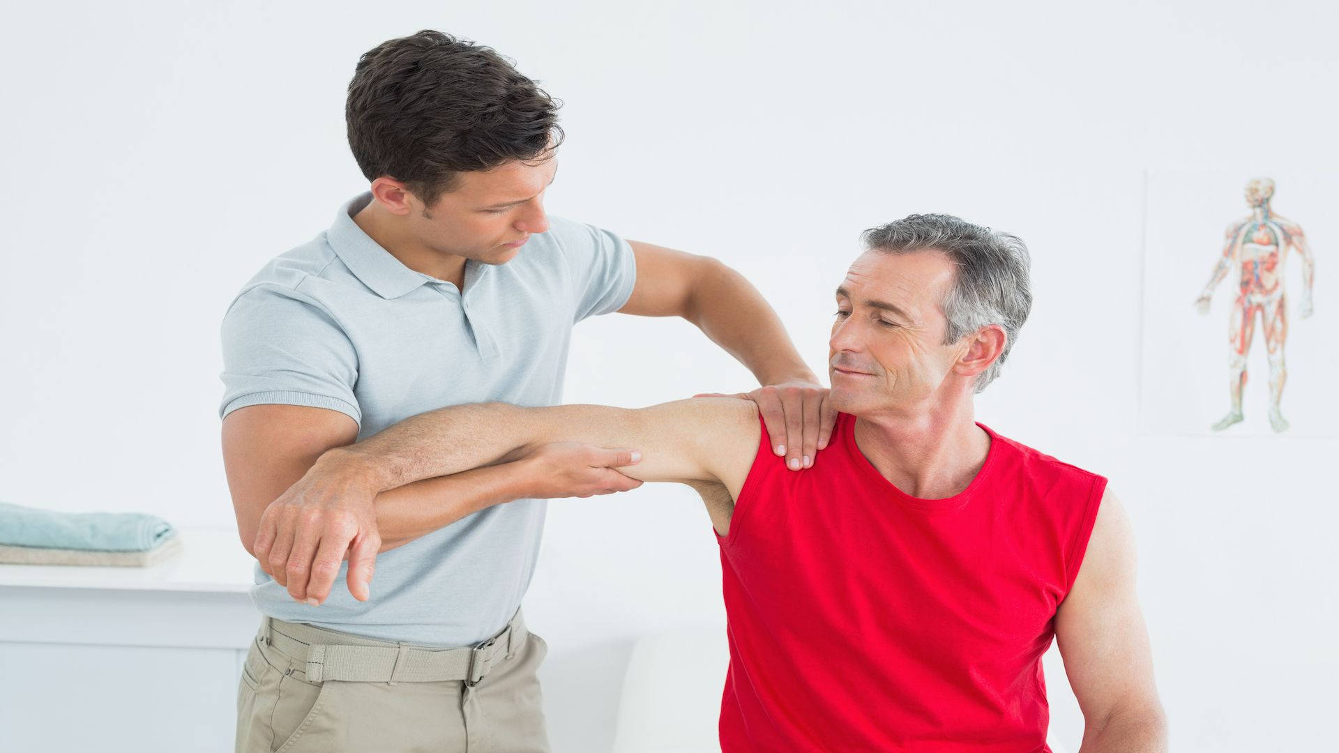 Massage 桌布 壁紙 — 一位身穿淺藍色襯衫的物理治療師，在明亮的診療室裡輕輕地操縱一位坐著的老年男性的手臂。