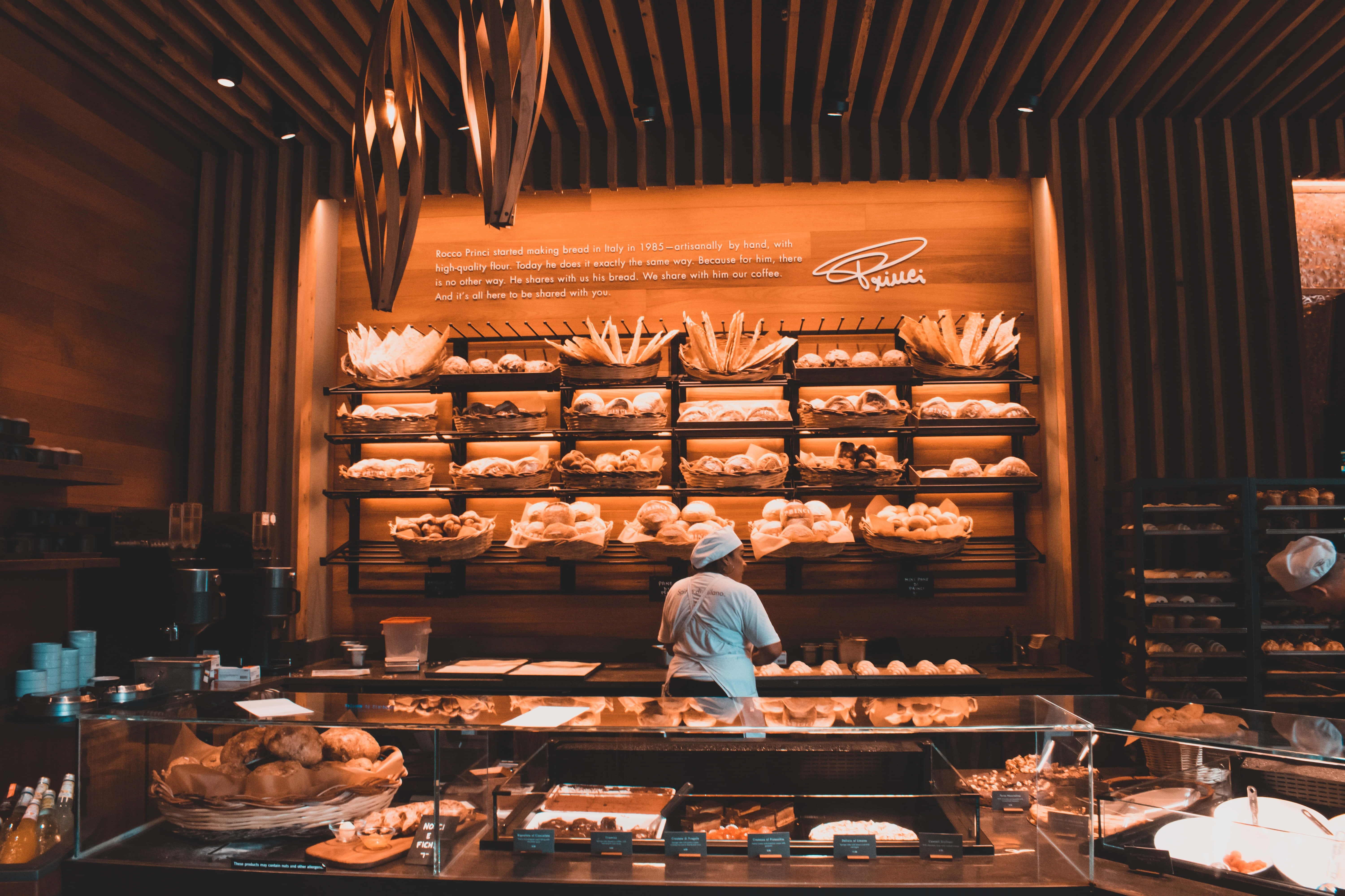 Bakery 桌布 壁紙 — 一位身穿淺藍色襯衫的麵包師傅在溫暖的木樑下，於一間有條紋牆壁和玻璃陳列櫃的舒適麵包店中擺放新鮮麵包。
