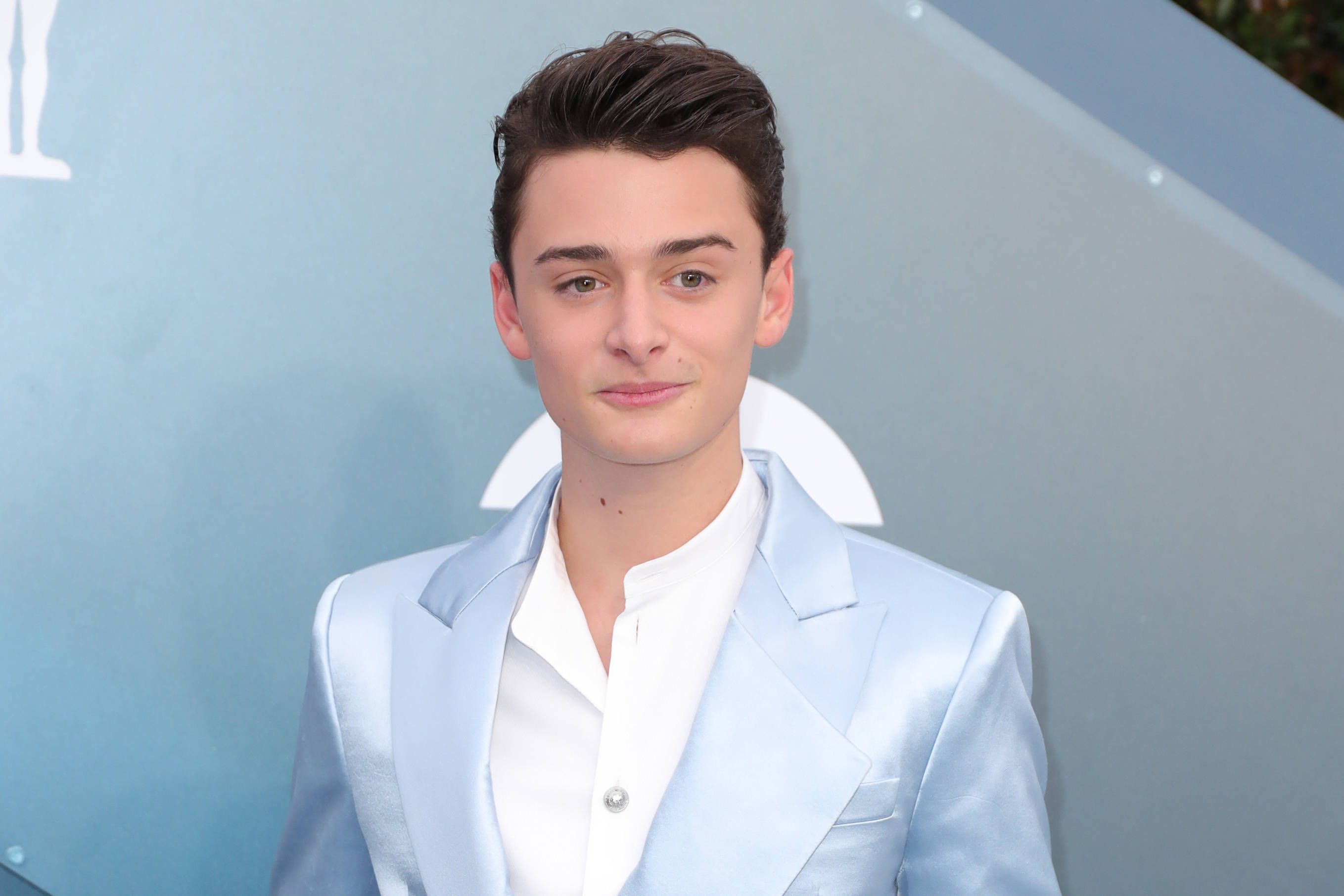 Celebrity 桌布 壁紙 — 一位身穿淺藍色西裝和白色襯衫的年輕男子，在帶有微妙白色標誌元素的柔和藍色背景前自信地擺姿勢。