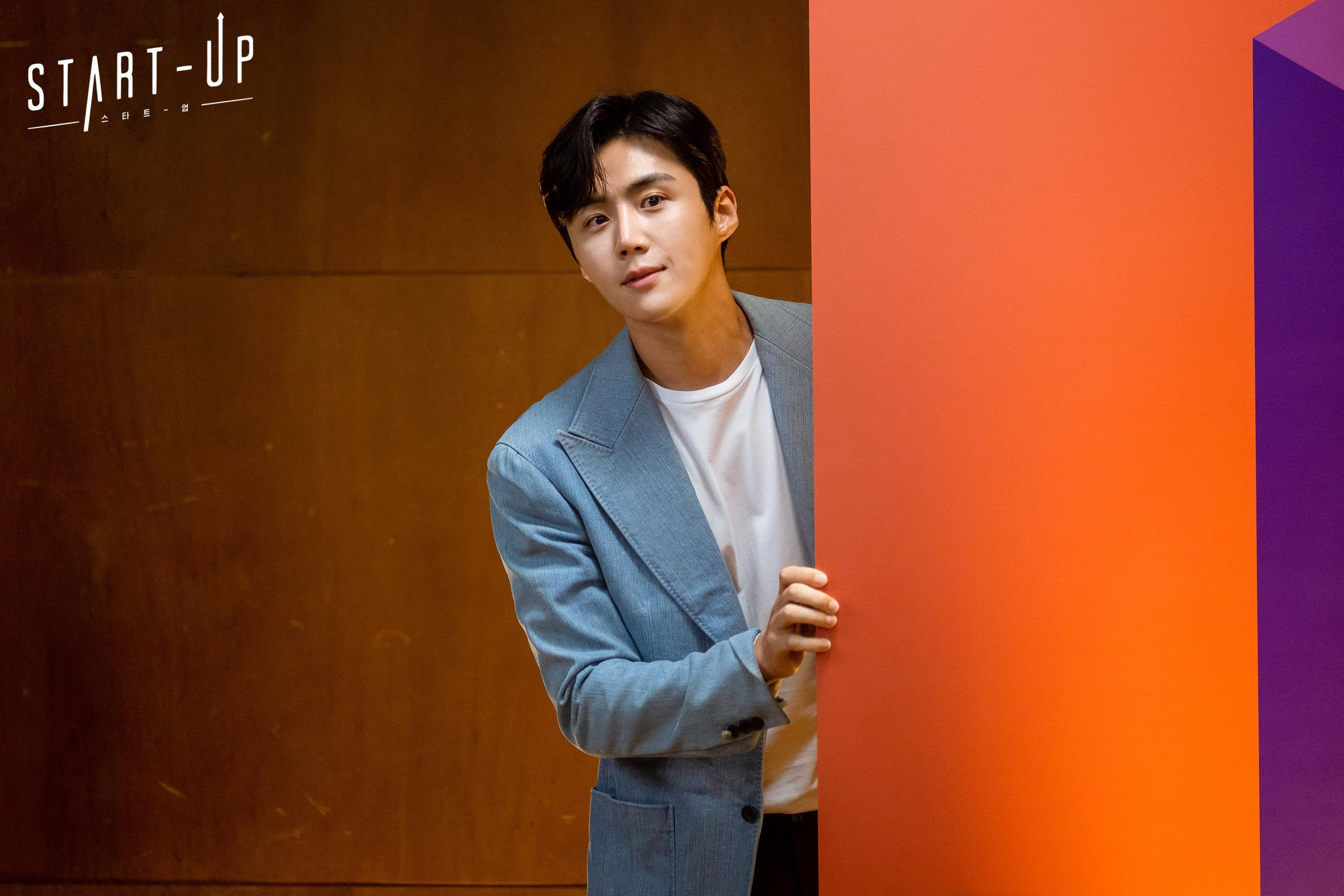 Kim Seon Ho 桌布 壁紙 — 一位身穿淺藍色西裝外套的年輕男子從充滿活力的橙色和紫色牆後探出頭來，背景是溫暖的木質面板。