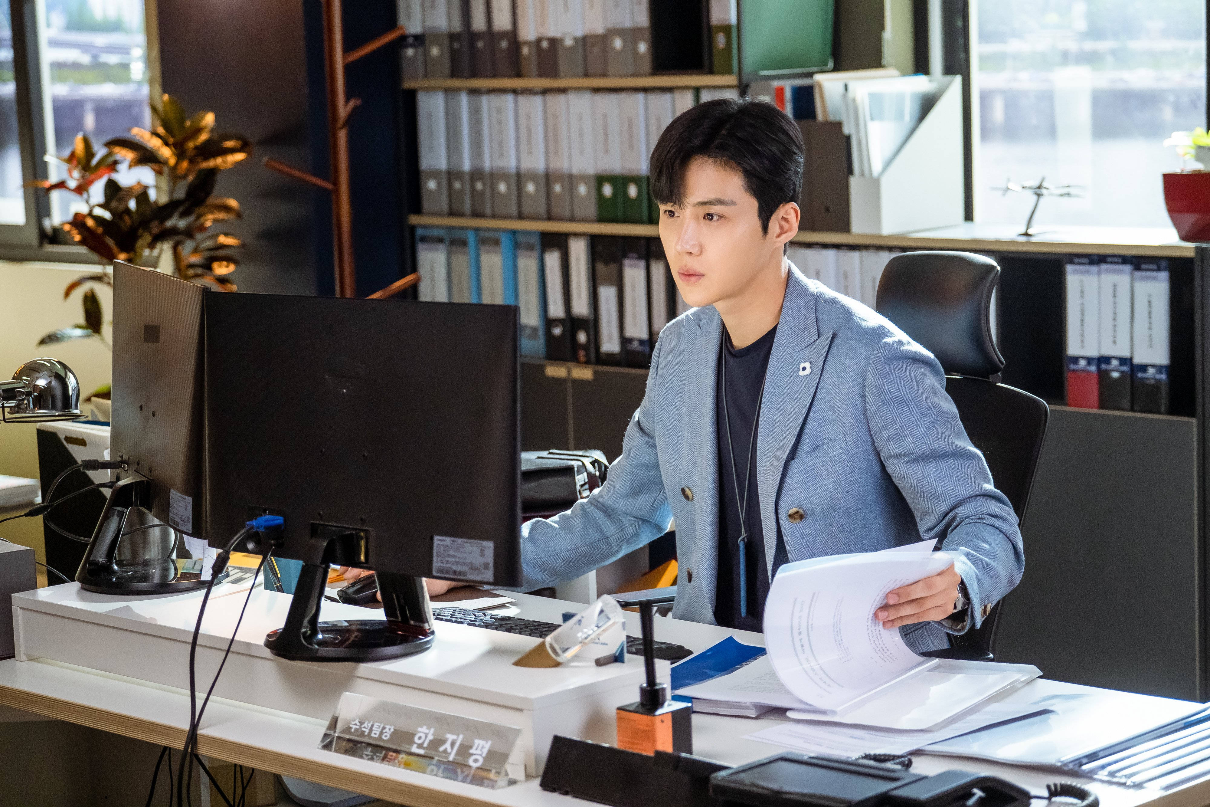 Kim Seon Ho 桌布 壁紙 — 一位身穿淺藍色西裝外套的年輕男子坐在雜亂的辦公桌前，在明亮的日光下審閱文件。