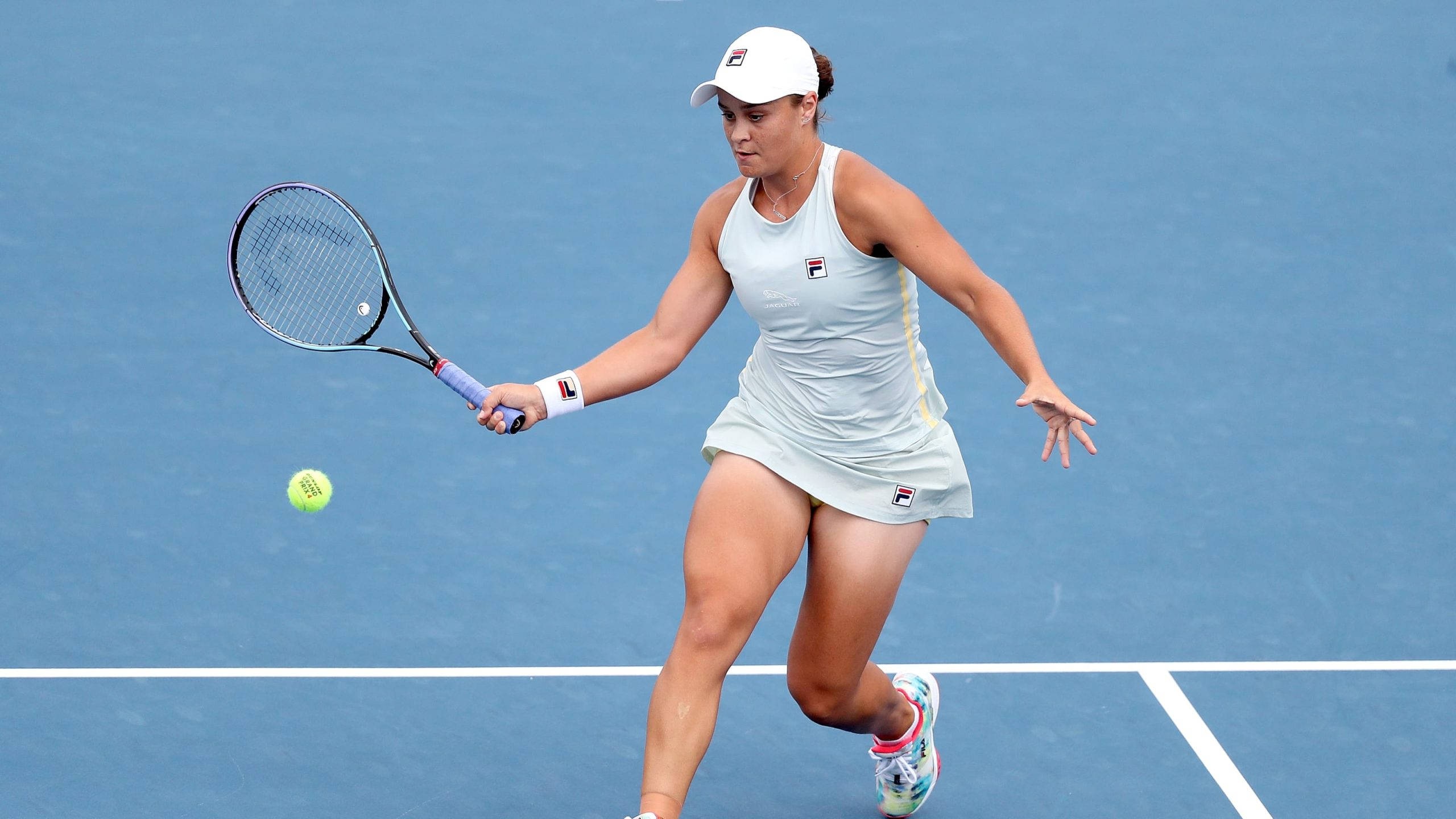 Ashleigh Barty 桌布 壁紙 — 一位身穿淺藍色連衣裙的女網球運動員在藍色硬地球場上向前猛衝，用球拍擊打一個亮黃色的網球。