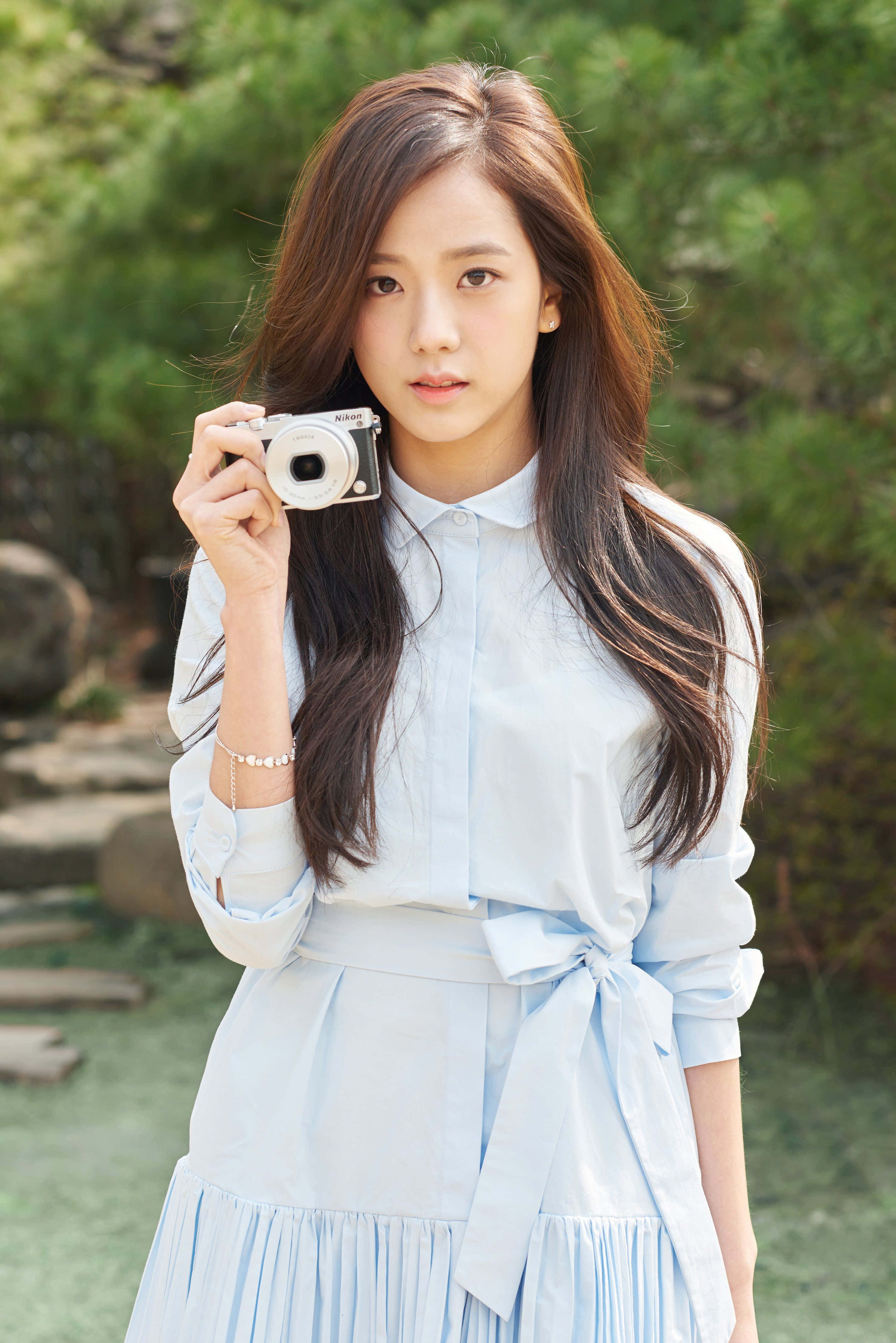 BLACKPINK 桌布 壁紙 — 一位身穿淺藍色連衣裙的年輕女子，在陽光普照的花園中，身後有綠色樹葉，手持一部銀色相機。