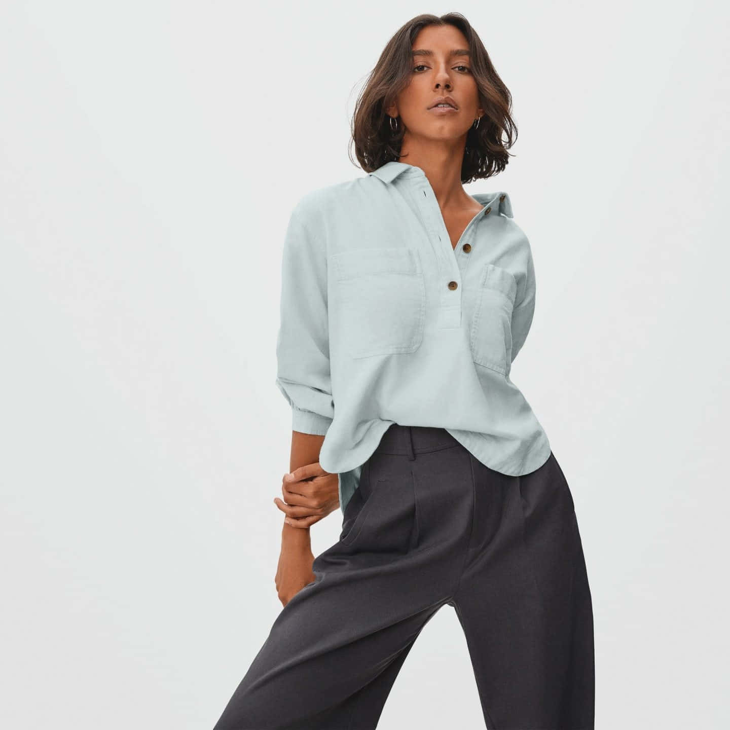 Minimalist Fashion 桌布 壁紙 — 一位身穿淺藍色鈕扣襯衫和深灰色長褲的女性，在純白色背景前自信地擺姿勢。
