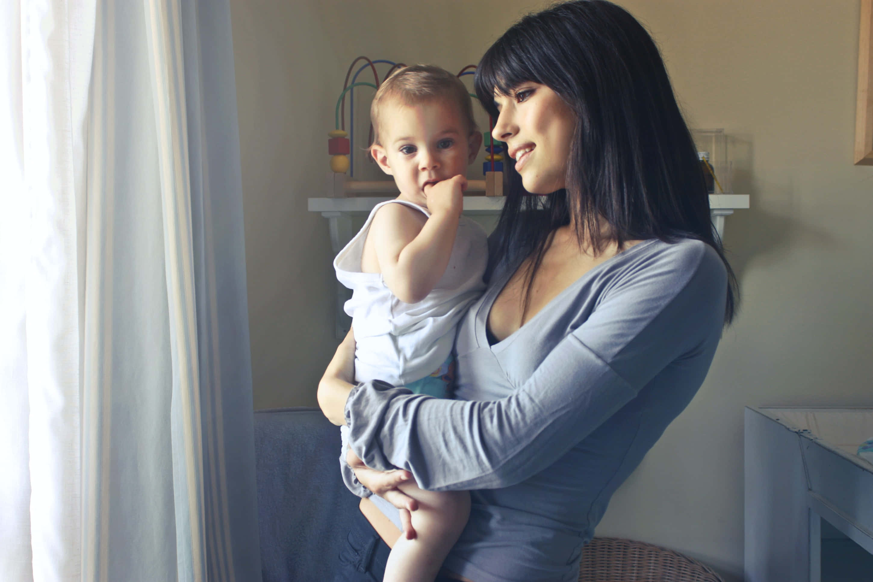 Mom 桌布 壁紙 — 一位身穿灰色上衣的女士，抱著身穿白色連身衣的嬰兒，坐在有薄紗窗簾的窗邊。