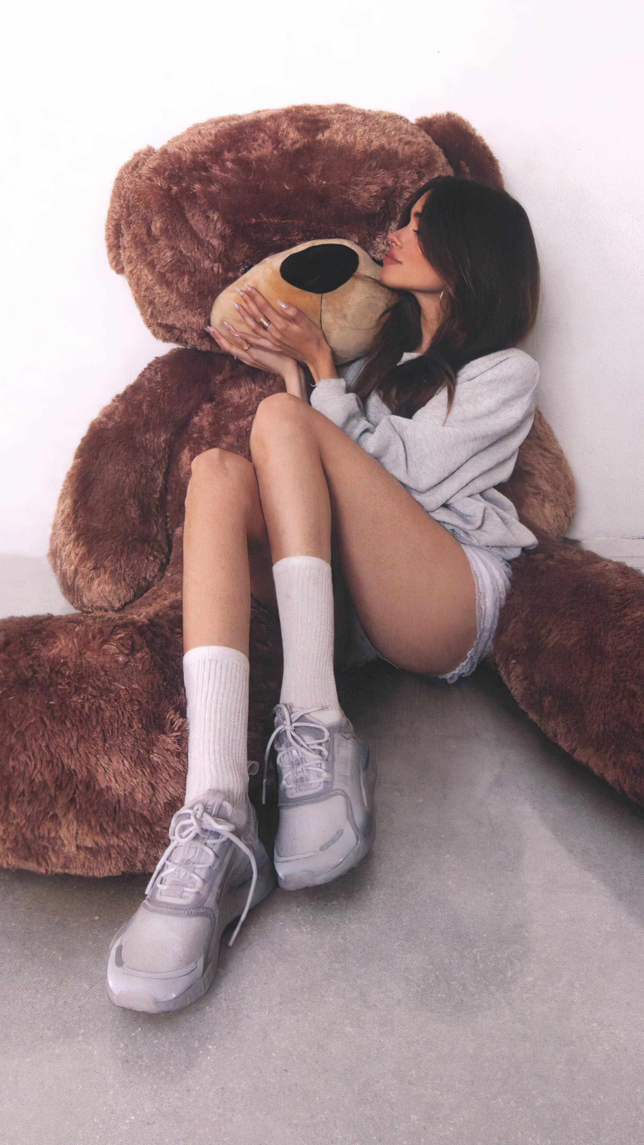 2160x3840 桌布 壁紙 — 一位身穿灰色休閒服的年輕女性坐在白色牆壁前的混凝土地板上，擁抱一隻巨大的棕色泰迪熊。