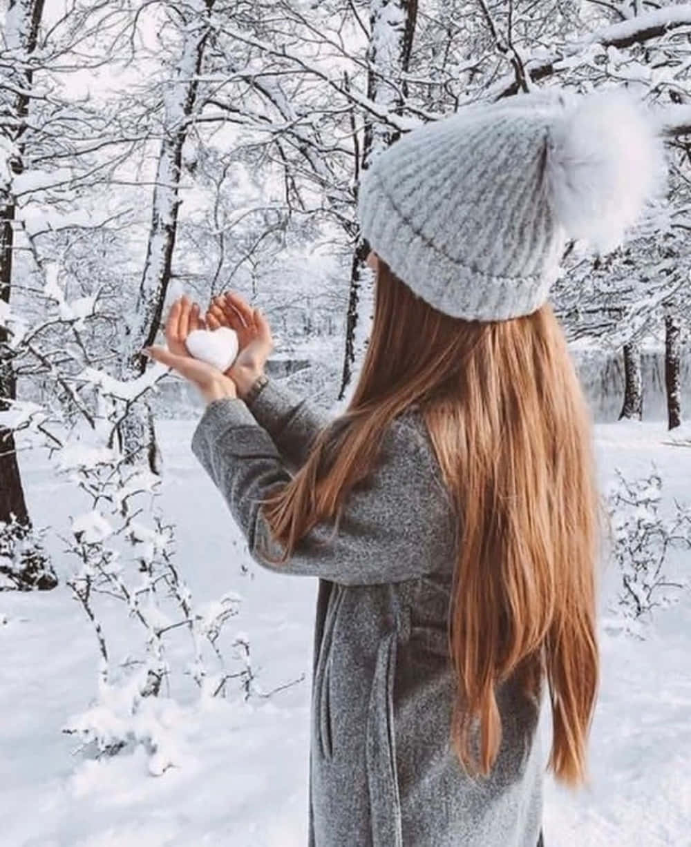 Winter Fashion 桌布 壁紙 — 一位身穿灰色外套和針織帽的女性在雪地森林中手持心形雪球。